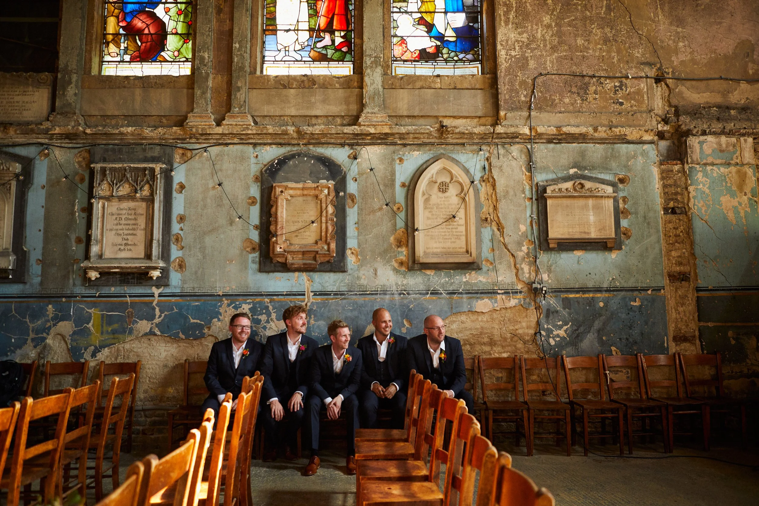 Five men sitting on a bench in an abandoned church with peeling paint and stained glass windows, dressed in formal suits with boutonnières, some smiling and chatting.