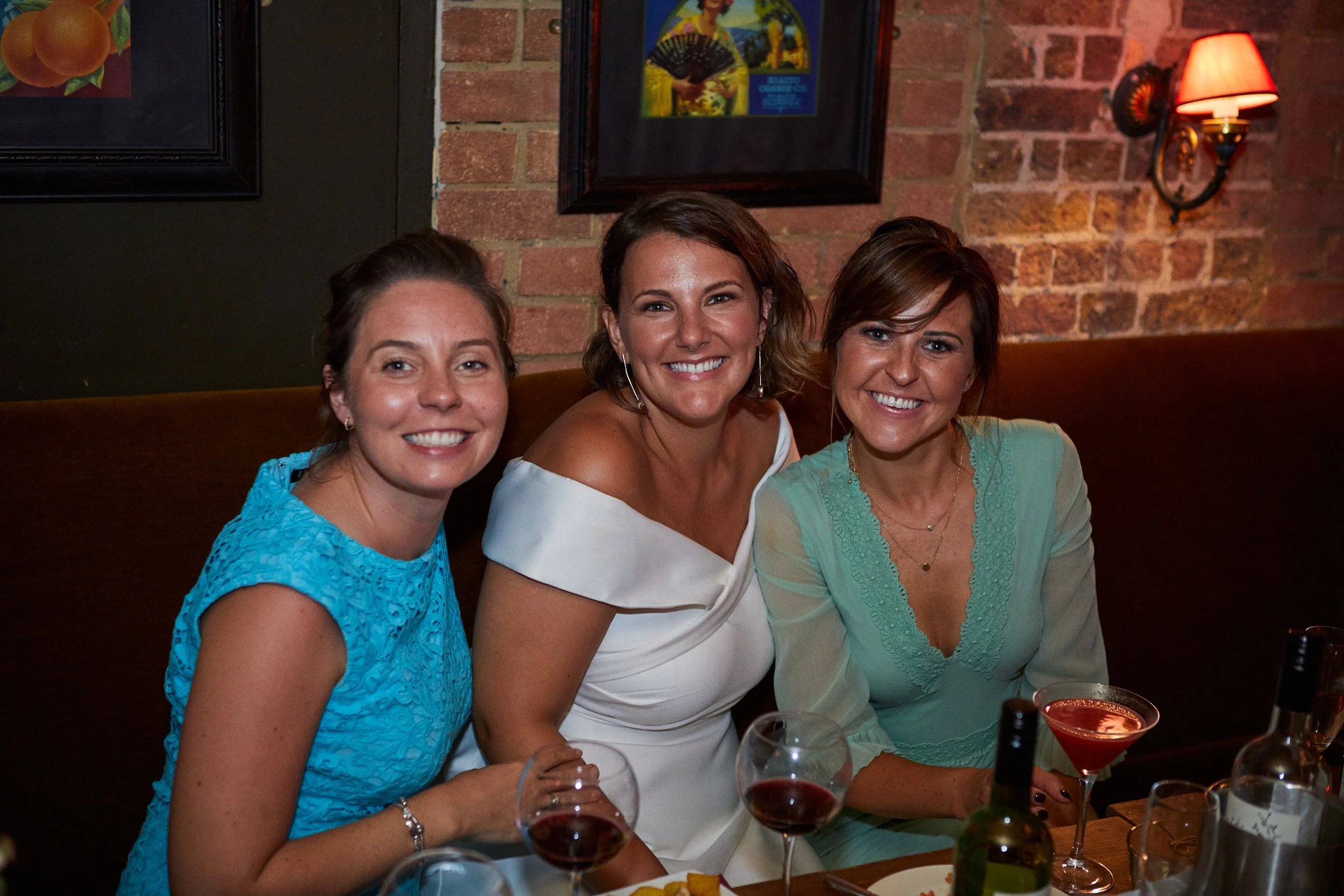 Three women sitting at a restaurant table, smiling for the photo, with drinks and bottles on the table. The background has brick walls and framed art, with a wall-mounted lamp.