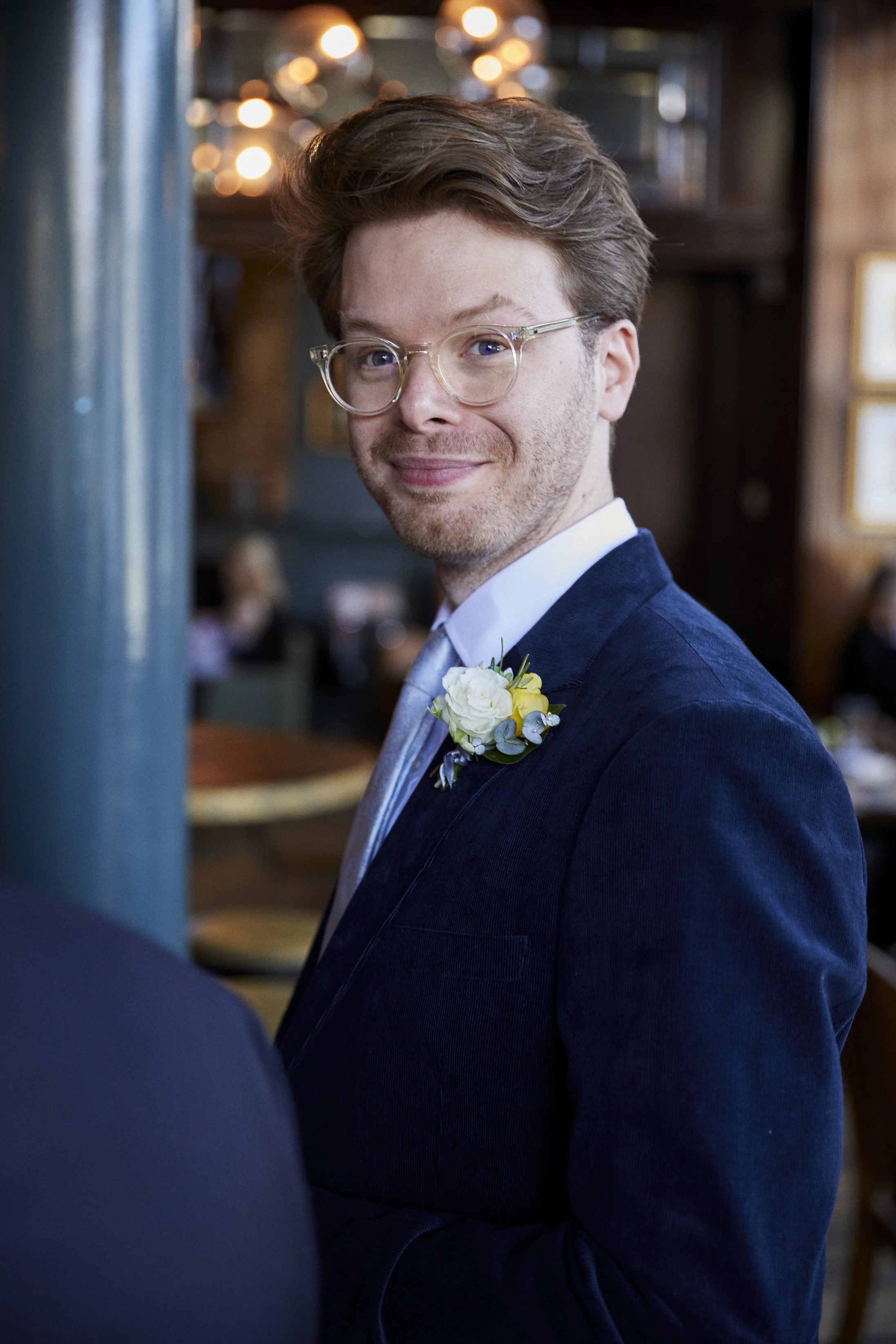 A man with glasses, smiling, wearing a dark blue suit jacket with a white shirt and a boutonniere, in a warmly lit indoor setting.