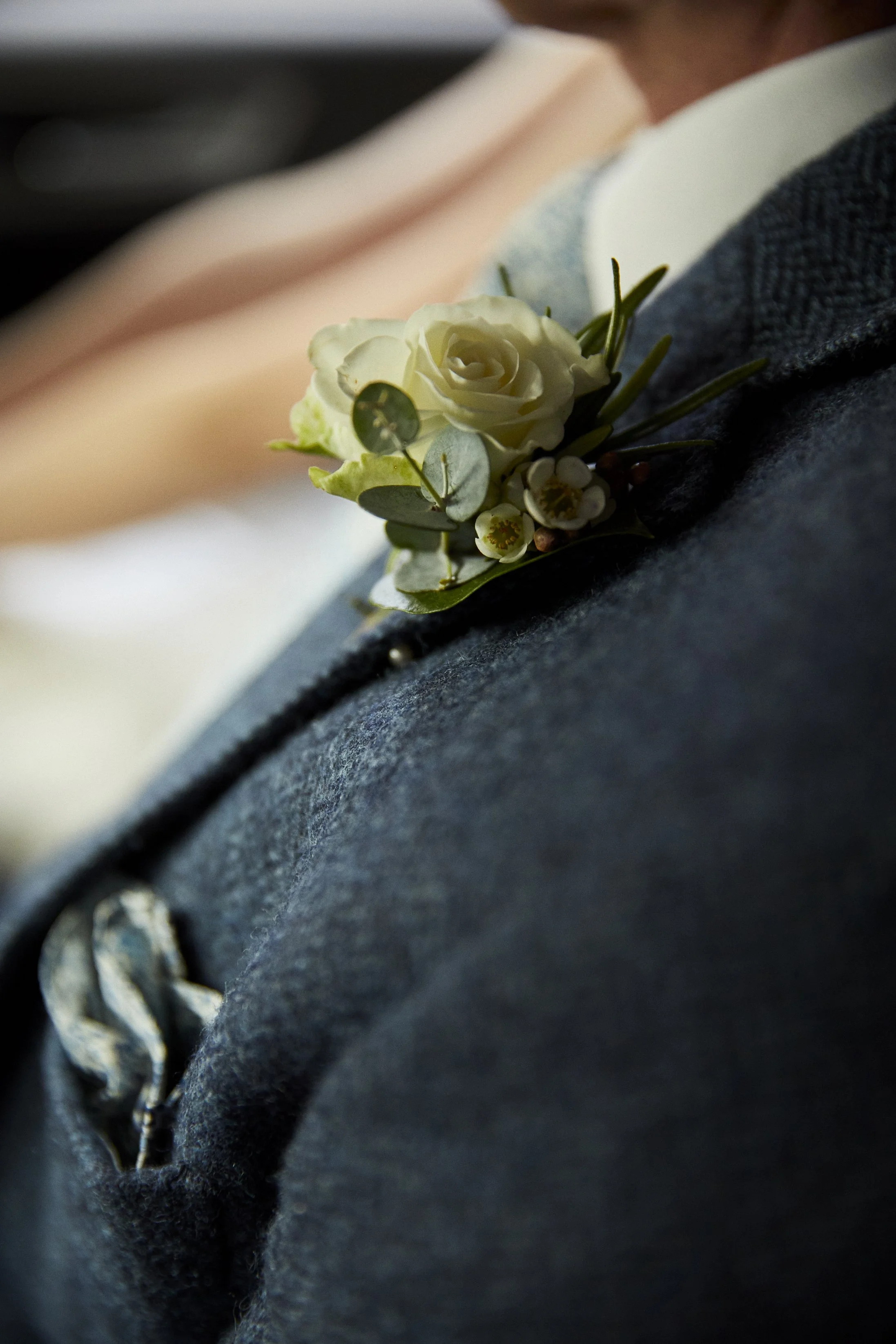 Close-up of a boutonniere with white roses and greenery pinned to a man's dark gray jacket.