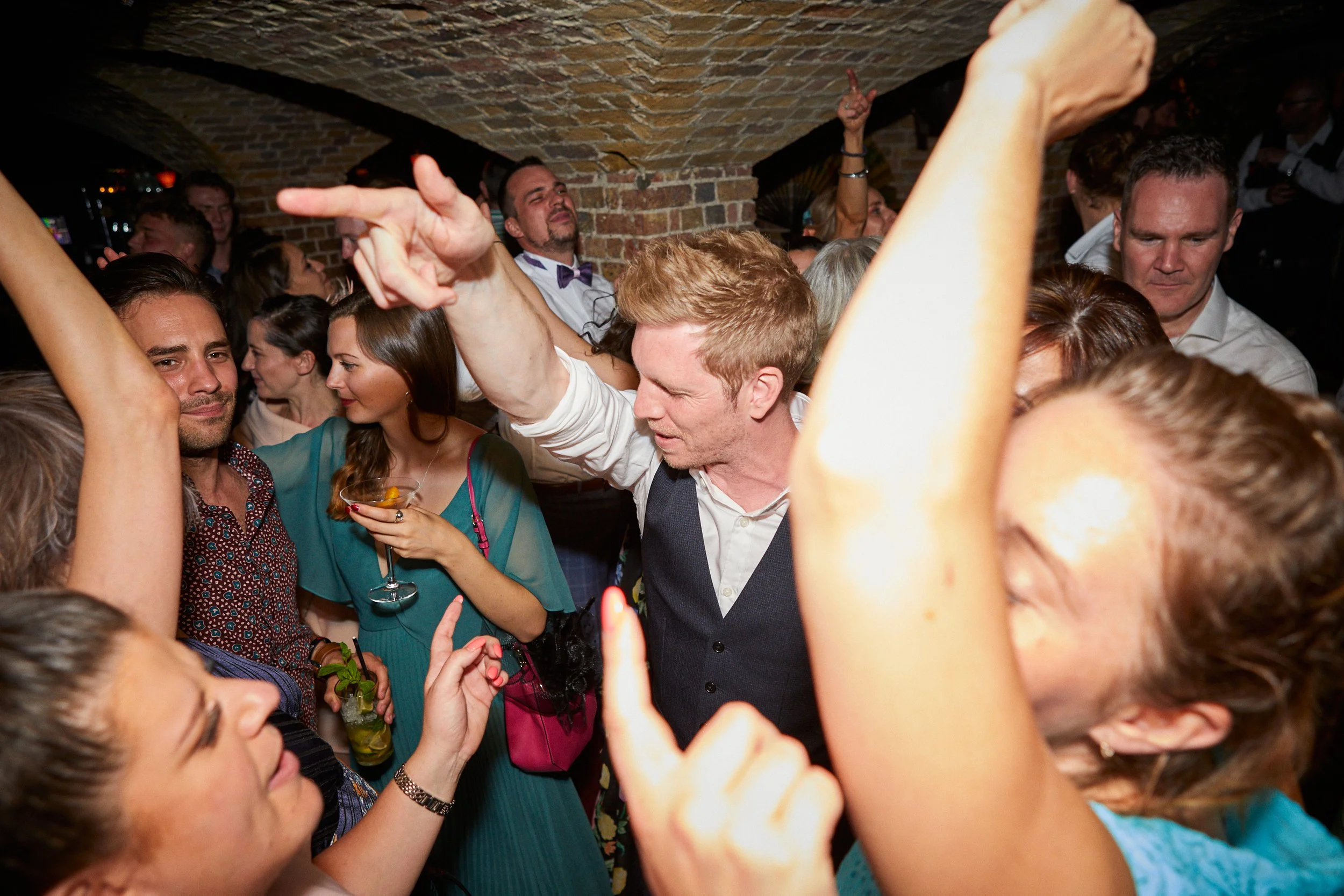 People dancing and socializing at a party in a dimly lit venue with a brick ceiling, enjoying drinks and music.