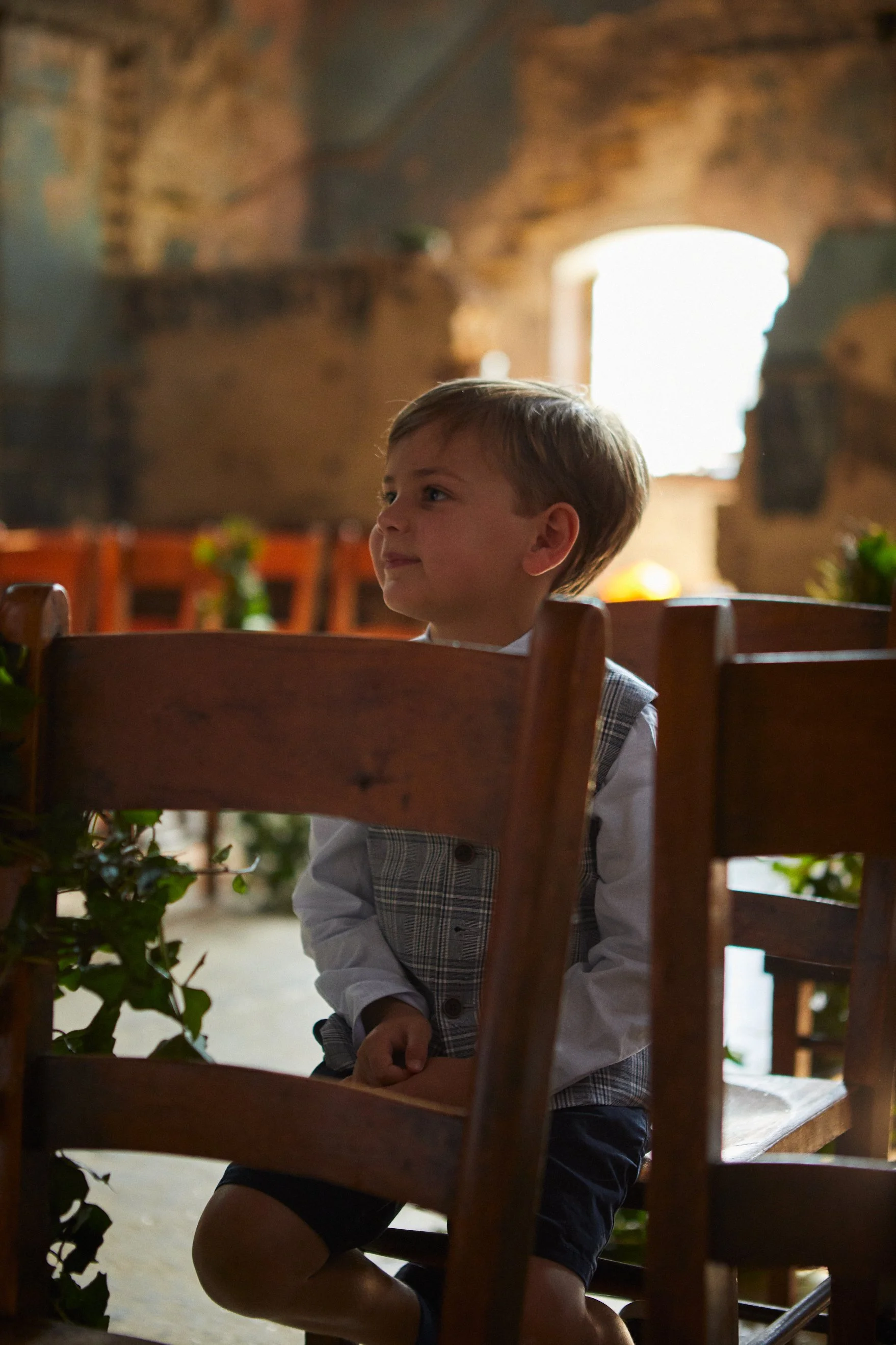 A young boy sitting indoors with wooden chairs and greenery, in a setting with exposed brick walls and a bright window in the background.