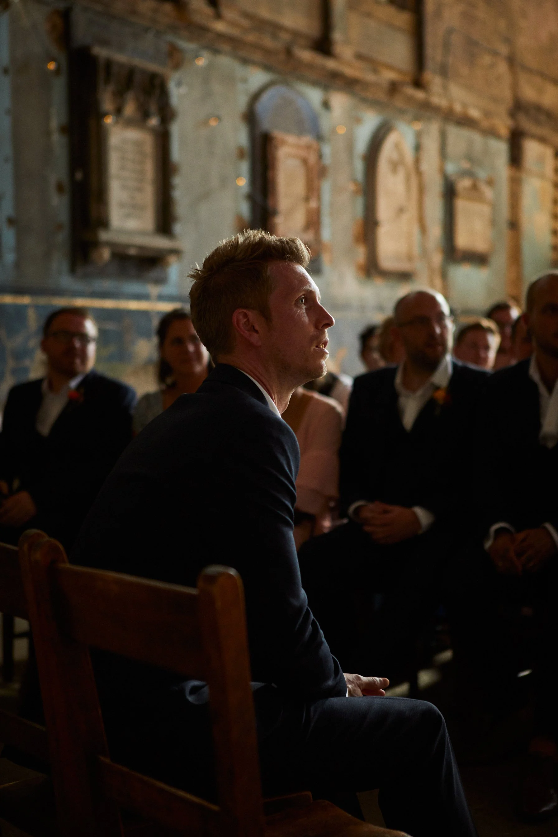 A man sitting in a dimly lit room with exposed brick and framed items on the wall, attending a formal event with other guests in the background.