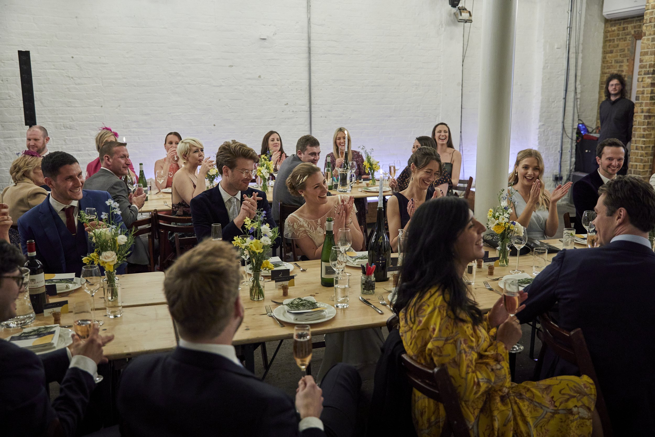 People at a wedding reception enjoying and clapping with flowers and wine glasses on the table, in a room with white brick walls.