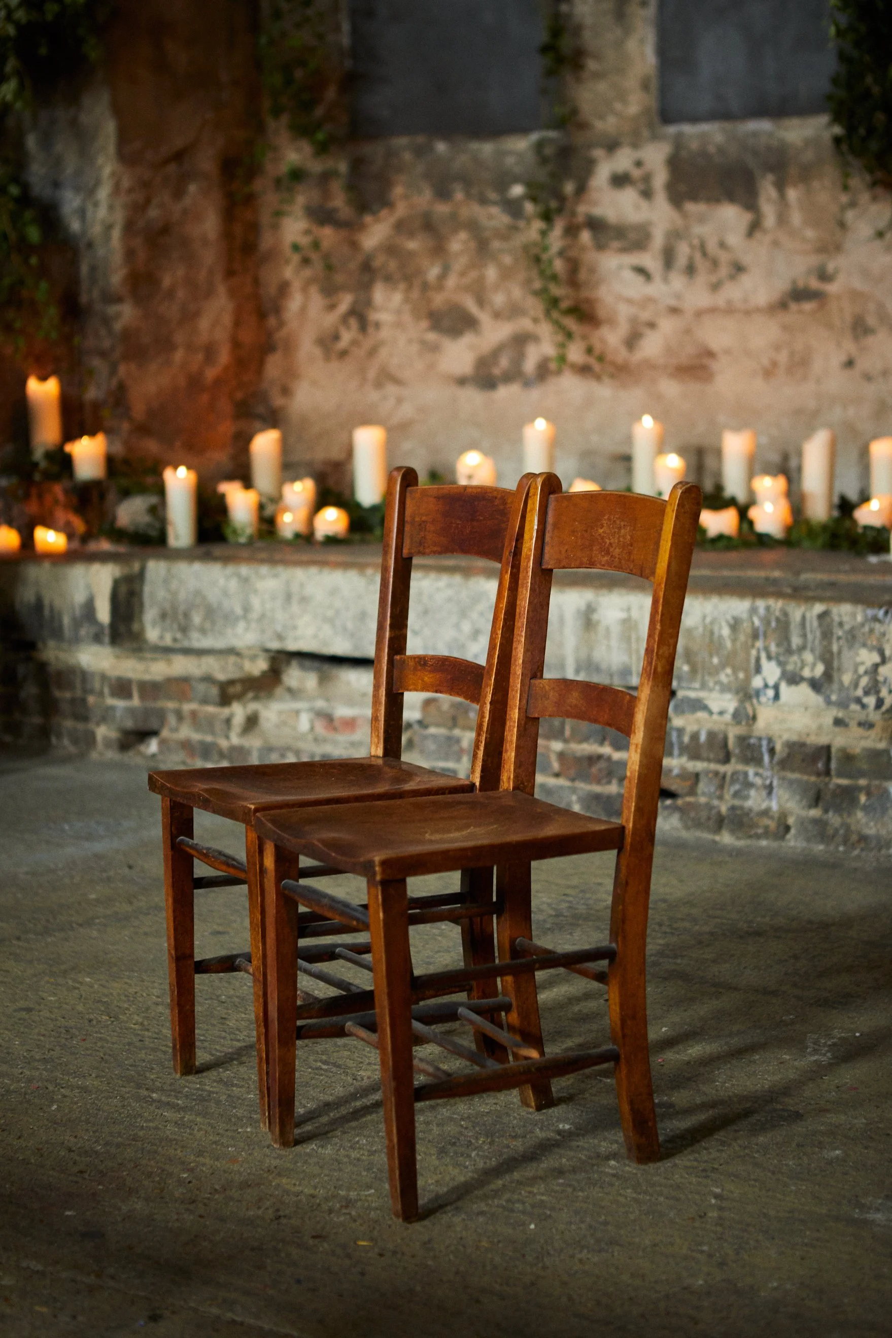 Two wooden chairs in front of a stone wall decorated with numerous lit candles.