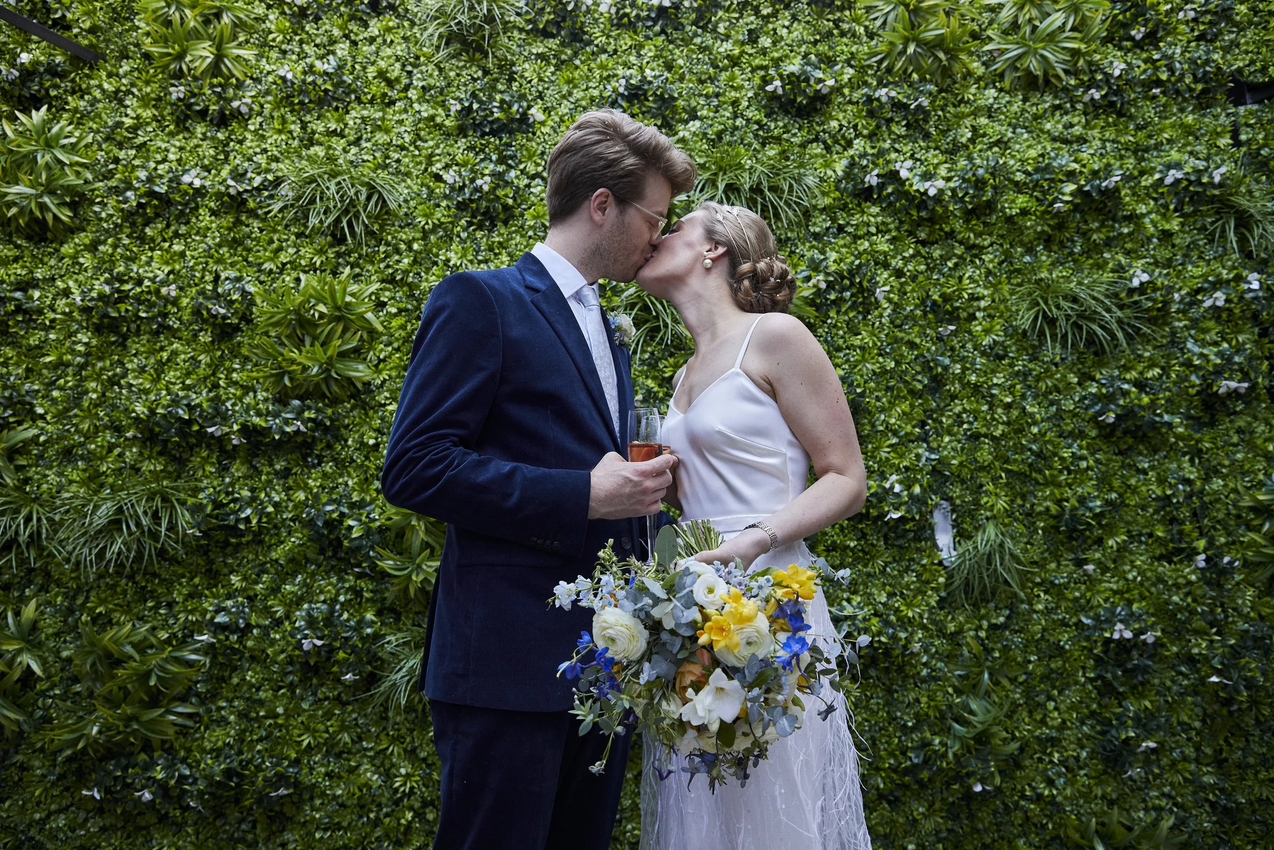 A bride and groom share a kiss at their wedding, with the groom holding a glass of champagne and the bride holding a bouquet of flowers, standing in front of a lush green hedge.