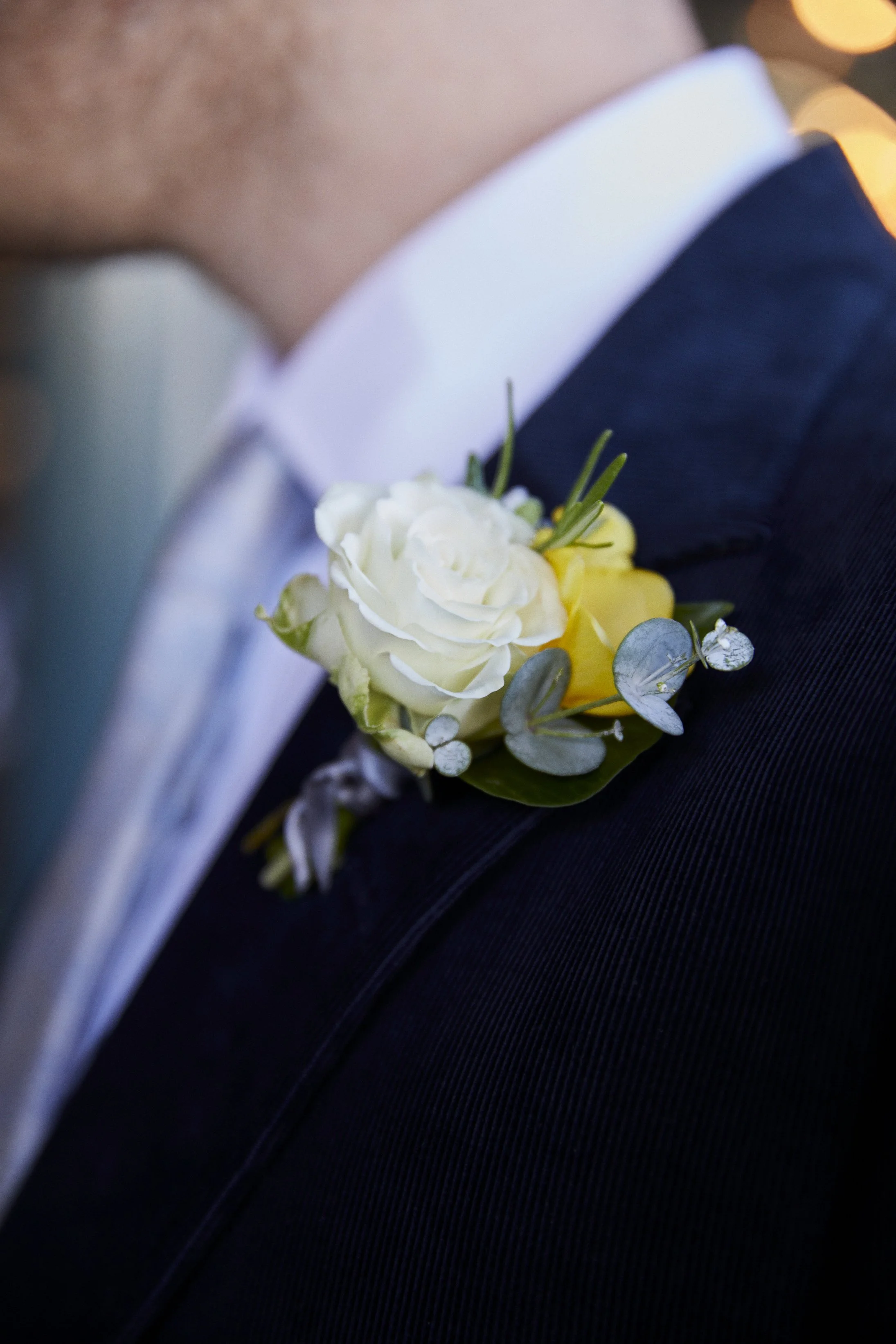 Close-up of a man's lapel with a white and pale yellow boutonniere floral arrangement pinned to it, featuring white roses, yellow flowers, eucalyptus leaves, and small white accent flowers.