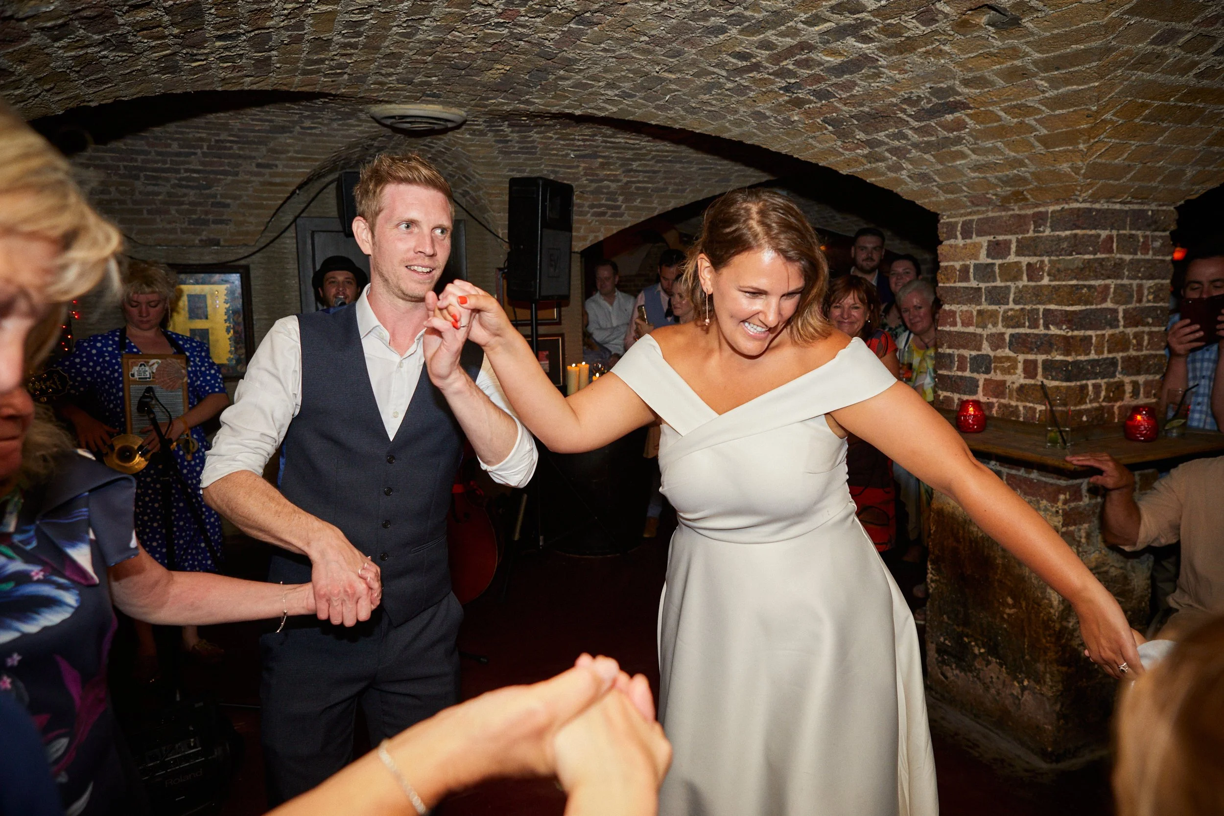 A bride and groom are dancing and holding hands at their wedding reception. The bride is wearing a white dress and the groom is wearing a vest and white shirt. Guests are observing and smiling in the background in a brick-walled venue.