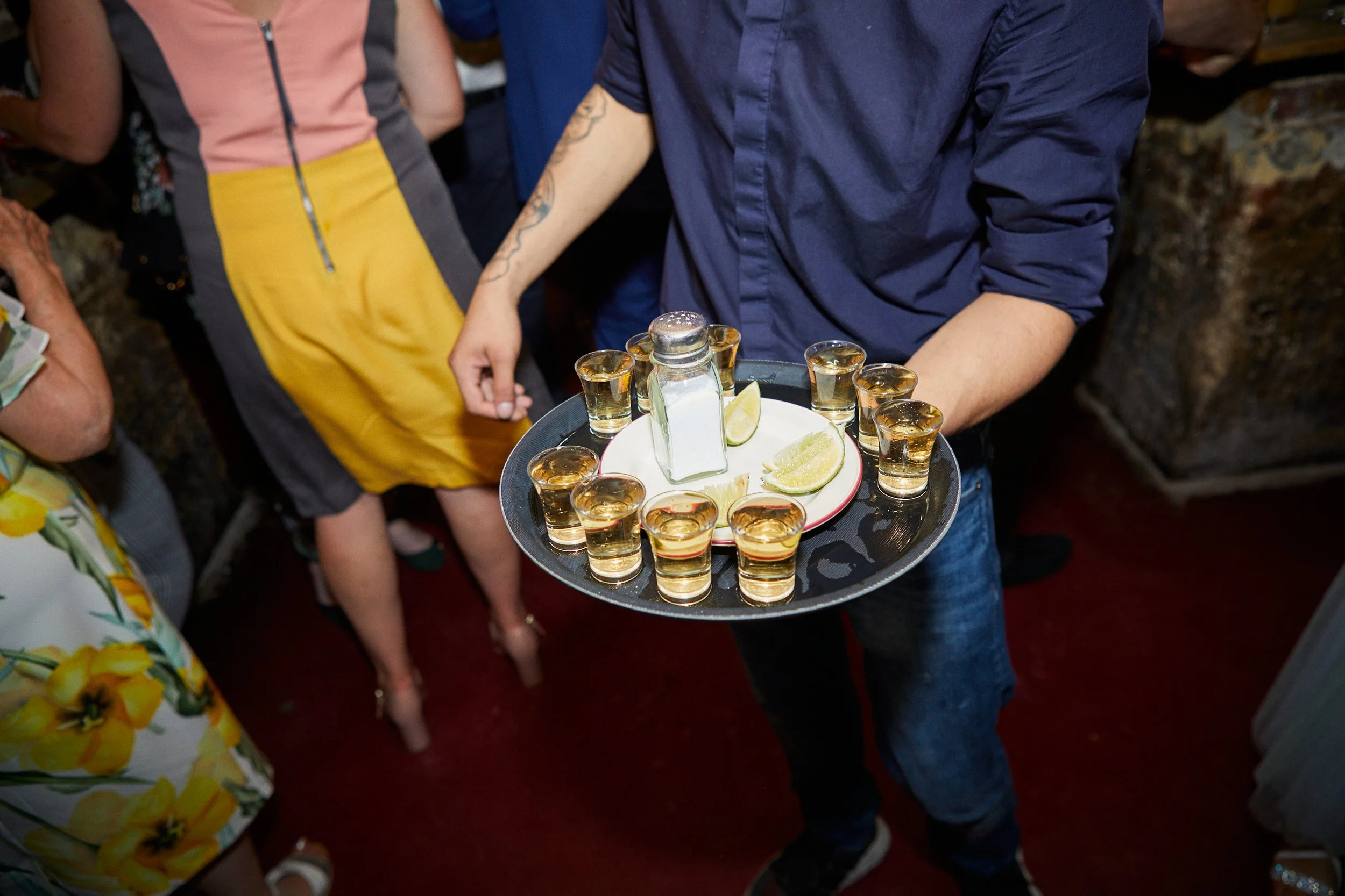 A waiter holding a black tray with small shot glasses of alcohol, lime wedges, and a salt shaker at a social gathering.