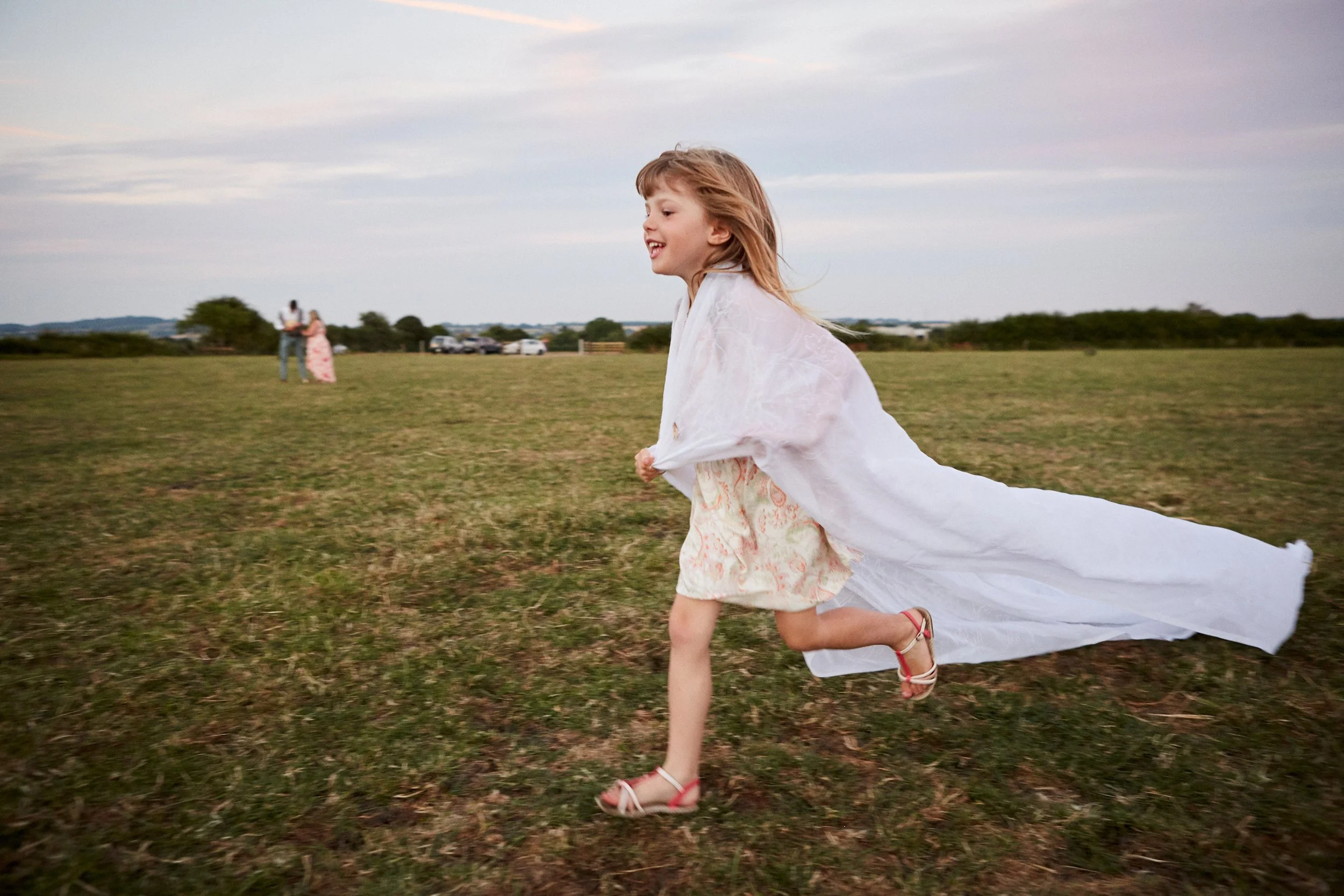 A young girl in a white dress and sandals running across an open grassy field during sunset, with two people in the background engaged in conversation.