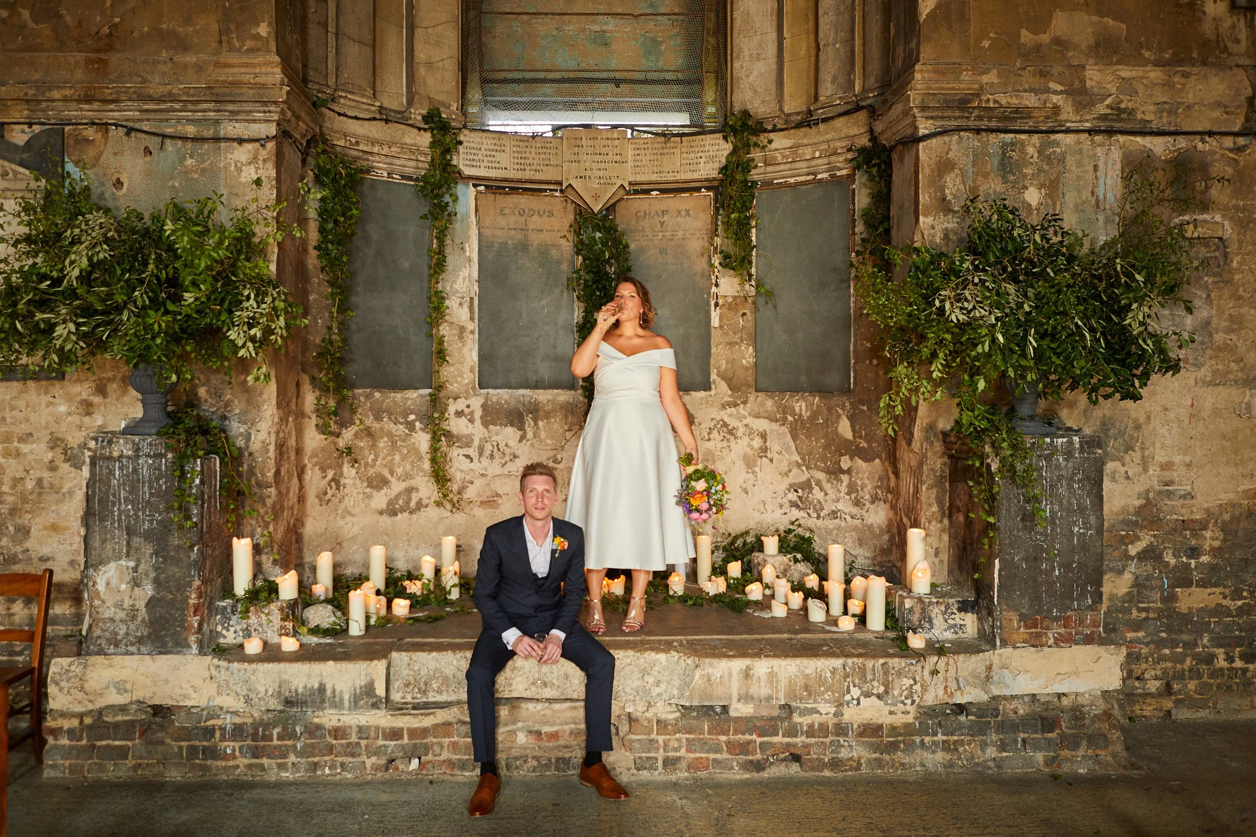 A newlywed couple in formal attire, the groom sitting on a brick ledge and the bride standing behind him, in an old rustic setting with candles and greenery decorations.