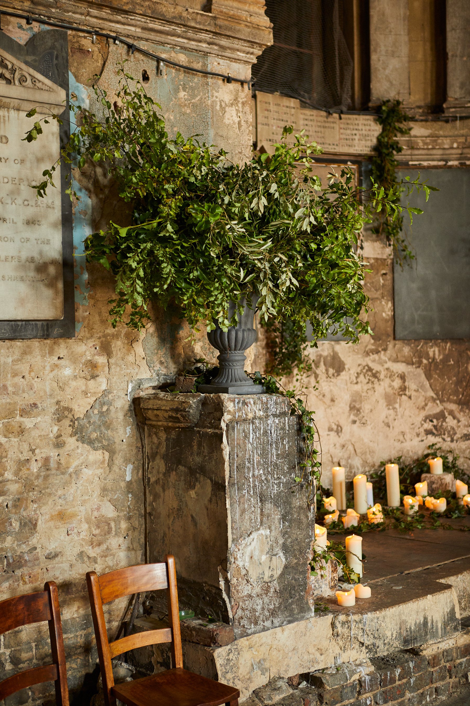 A large green leafy plant in a decorative gray vase sits on a stone pedestal against a distressed brick wall, with dozens of lit candles on the ground nearby.