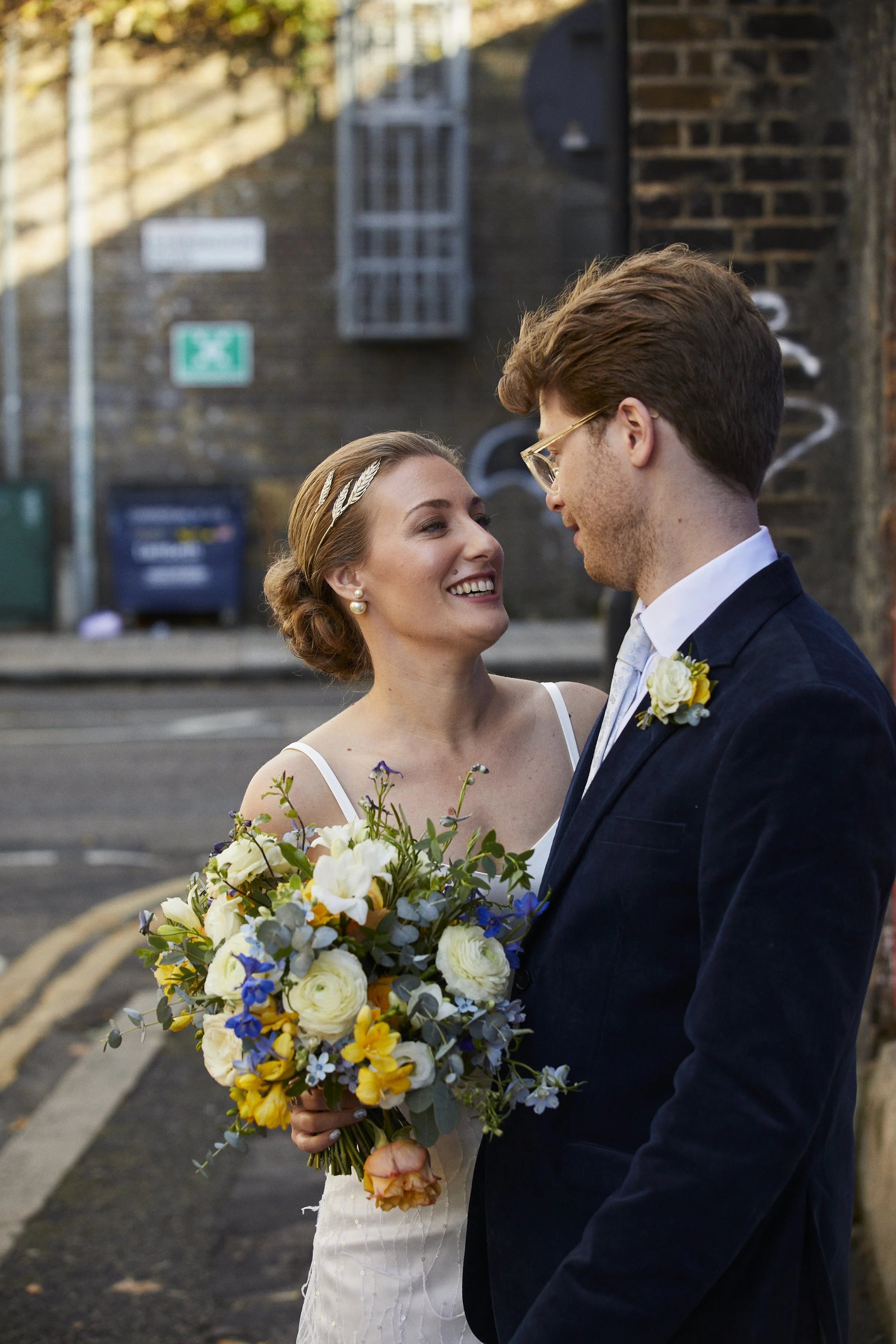 A bride and groom are standing close together outdoors, smiling and looking at each other. The bride is holding a bouquet of colorful flowers, and the groom is wearing glasses and a dark suit with a boutonniere. The background features a brick wall a