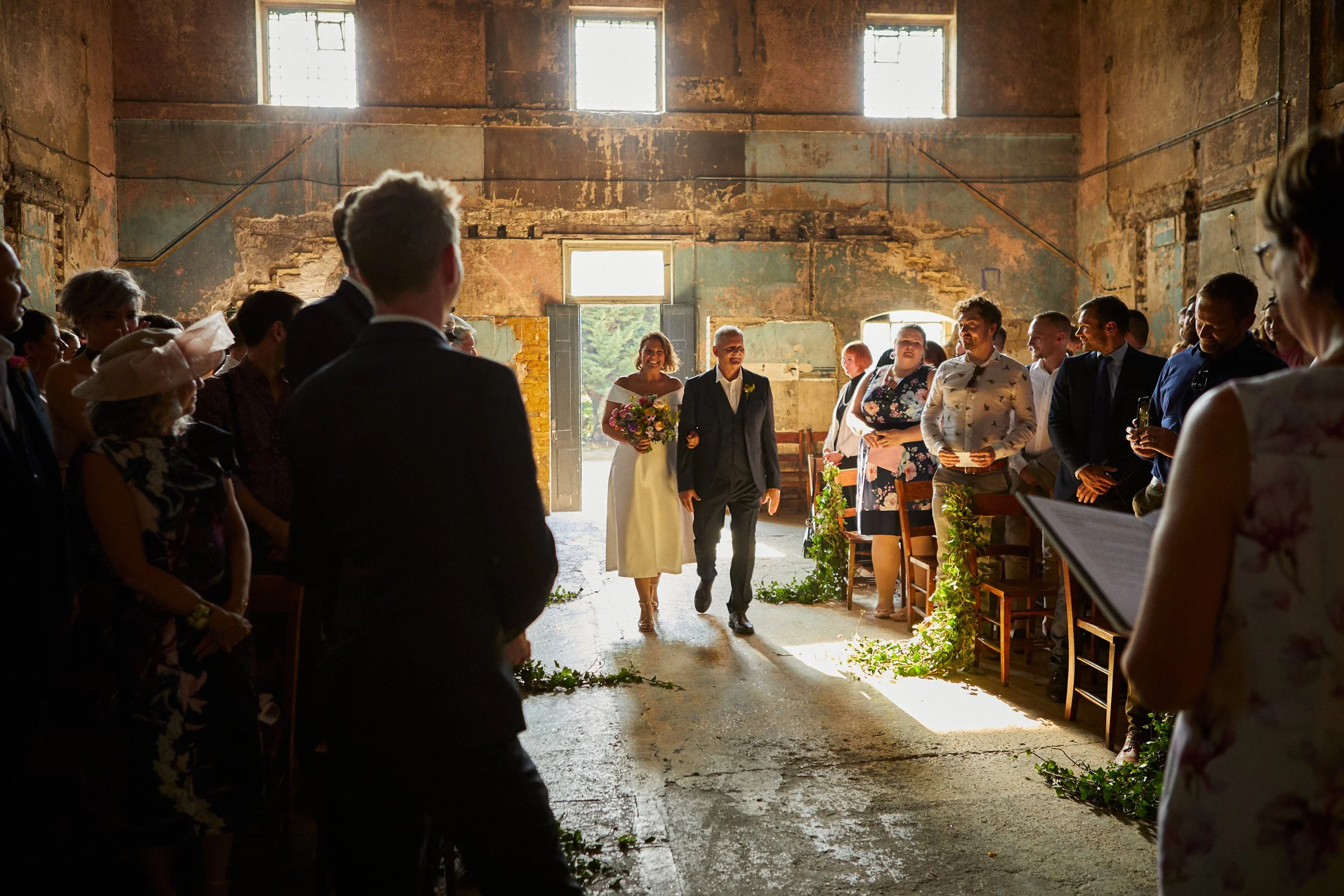 People attending a wedding ceremony inside a rustic, weathered barn, with the bride walking down the aisle toward the groom, who is standing in front of the seated guests, all illuminated by natural light coming through the windows.