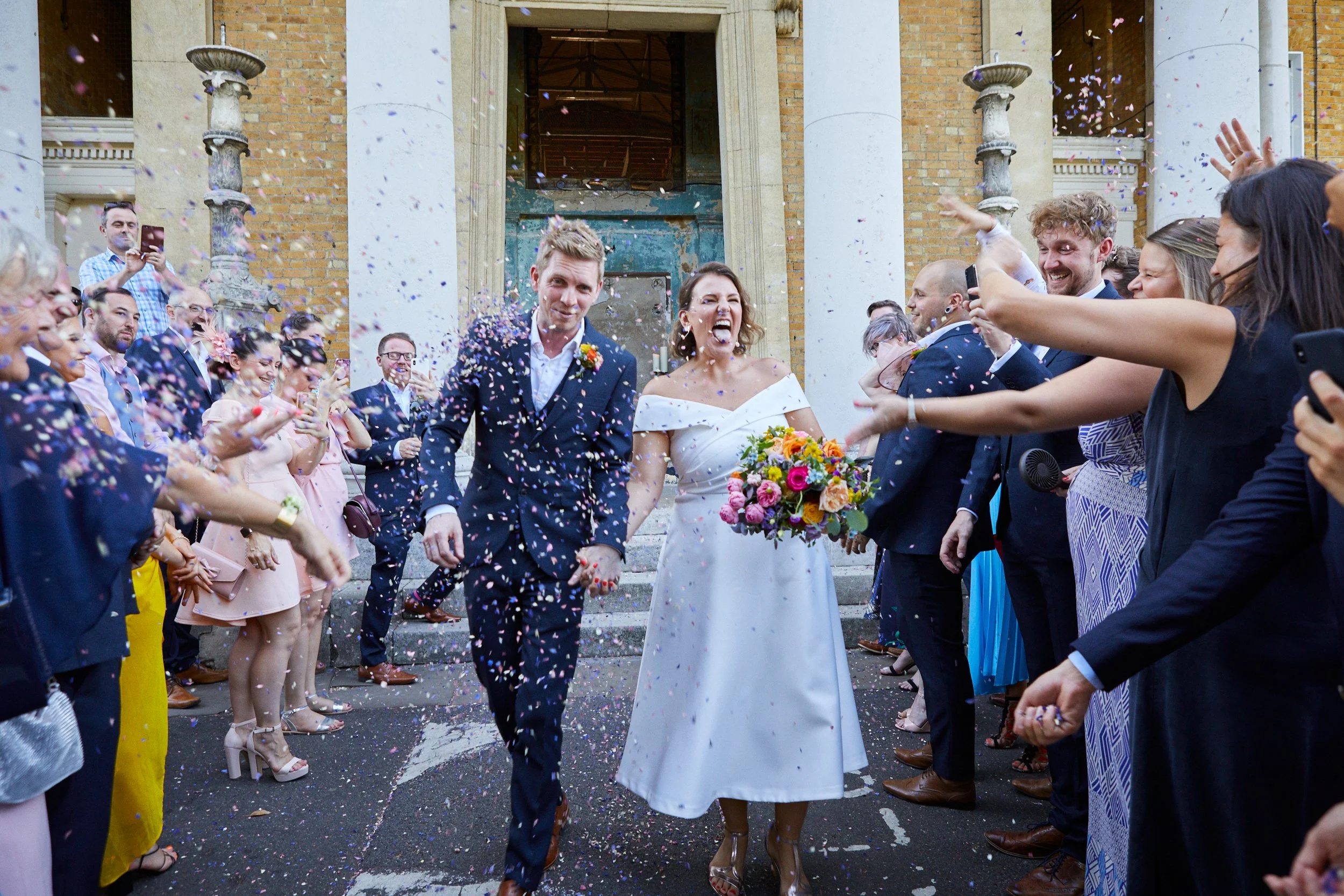 A newlywed couple walking hand-in-hand through a crowd of celebrating wedding guests, with confetti falling around them in front of a brick building with white columns.