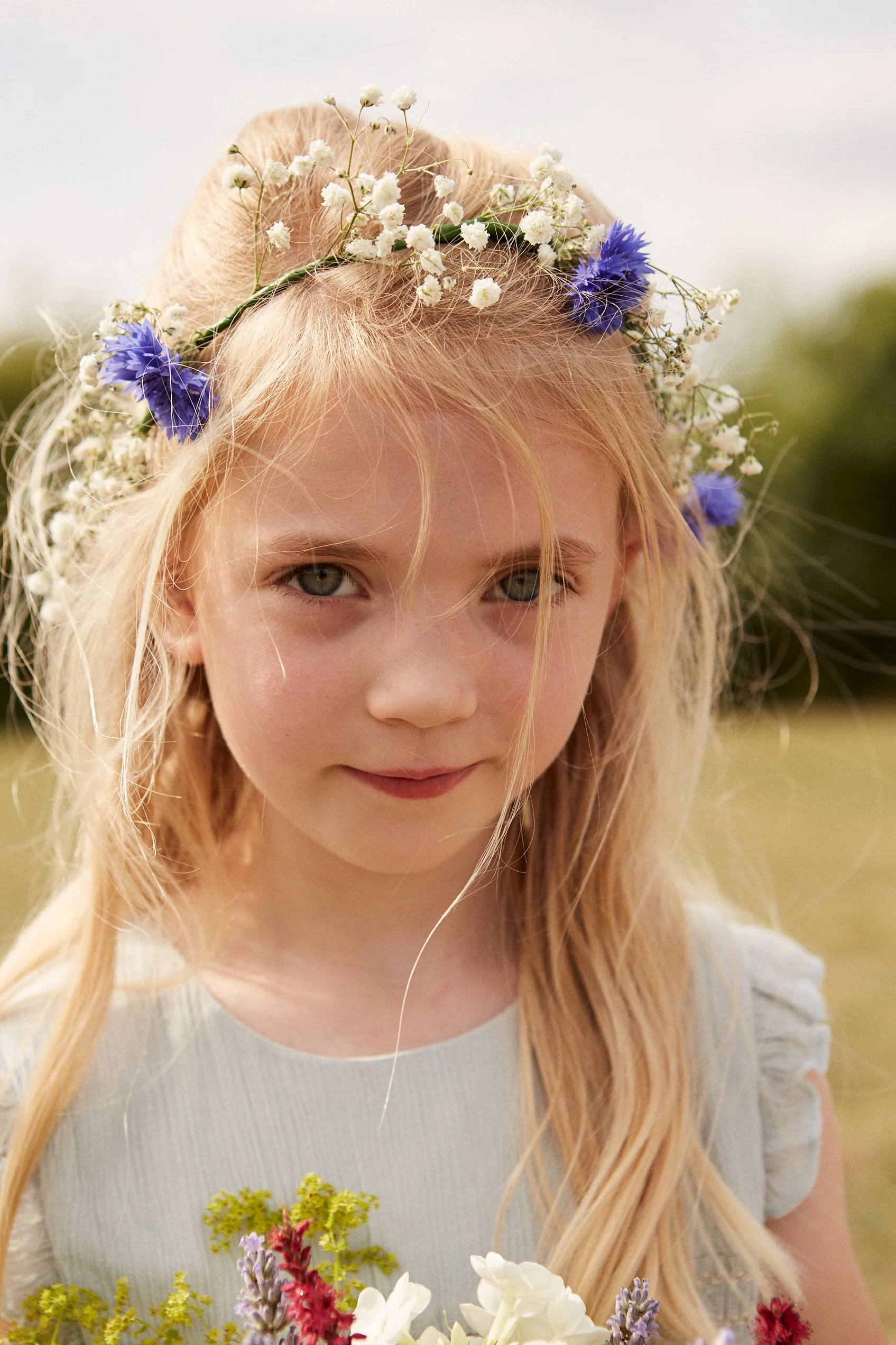 A young girl with blonde hair wearing a flower crown of white baby's breath and purple flowers, holding a bouquet of mixed flowers, outdoors.