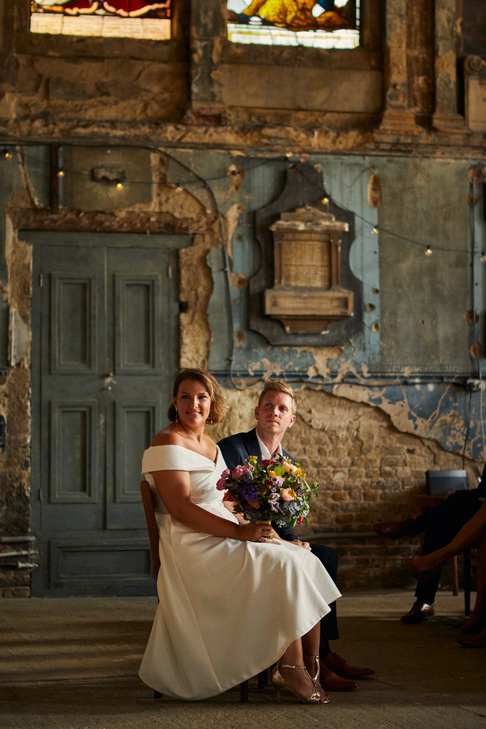 A bride and groom seated during their wedding ceremony in a rustic venue with exposed brick and wooden beams. The bride is holding a colorful bouquet, and both are dressed formally.