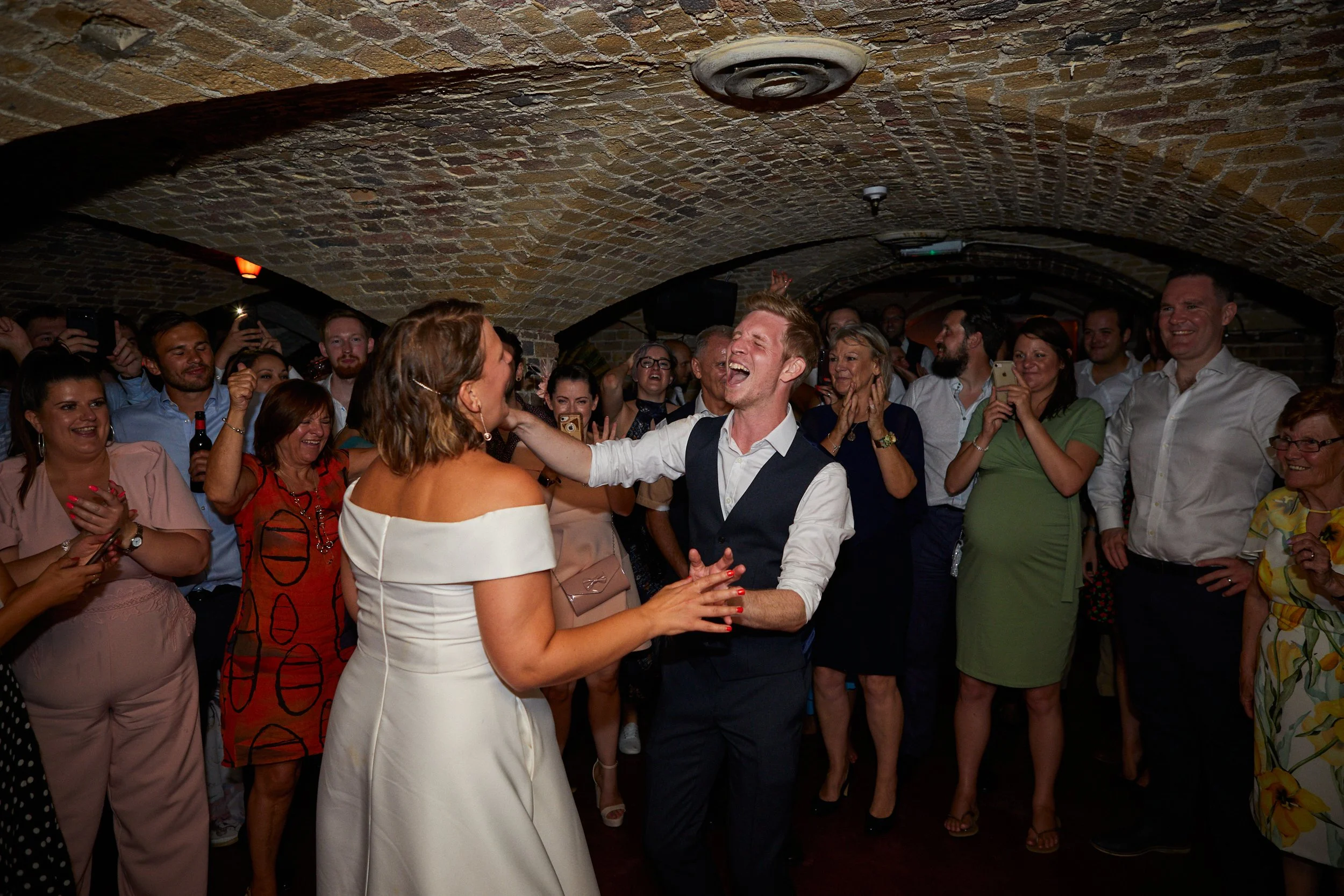 A bride and groom dancing joyfully at their wedding reception, surrounded by friends and family in an underground brick venue.