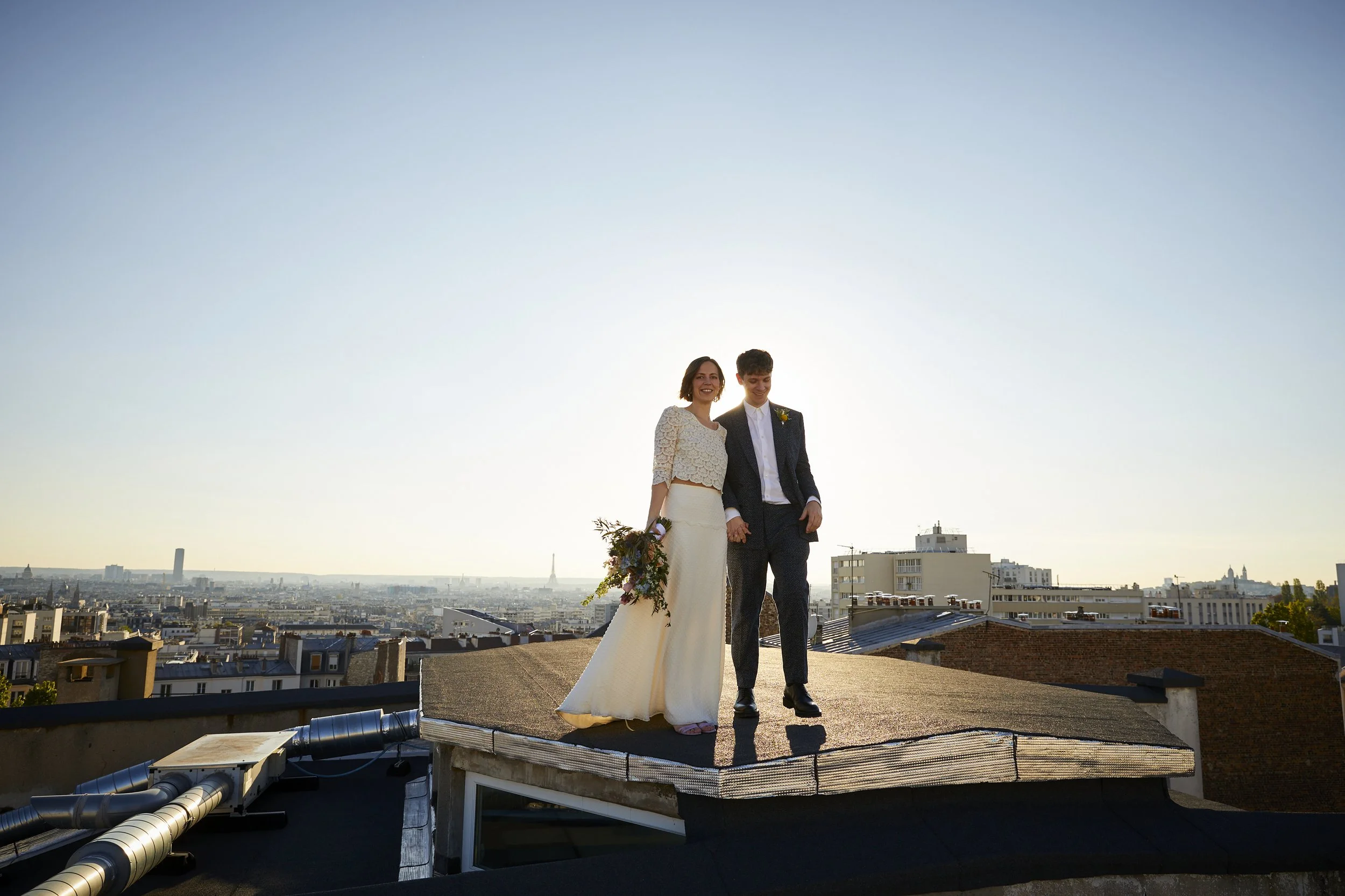 A couple in wedding attire standing on a rooftop with a city skyline in the background during sunset.