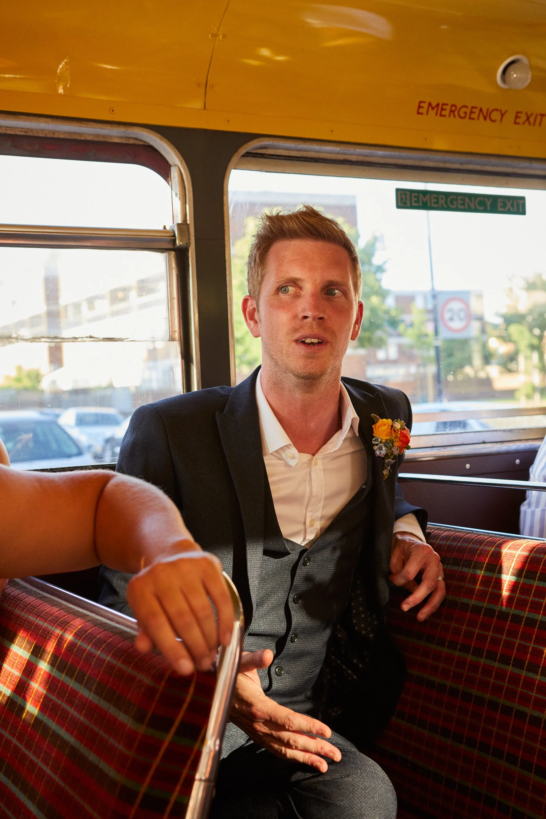A man wearing a suit with a flower boutonniere sitting on a patterned bus seat, inside a bus, with windows and an 'Emergency Exit' sign visible behind him.