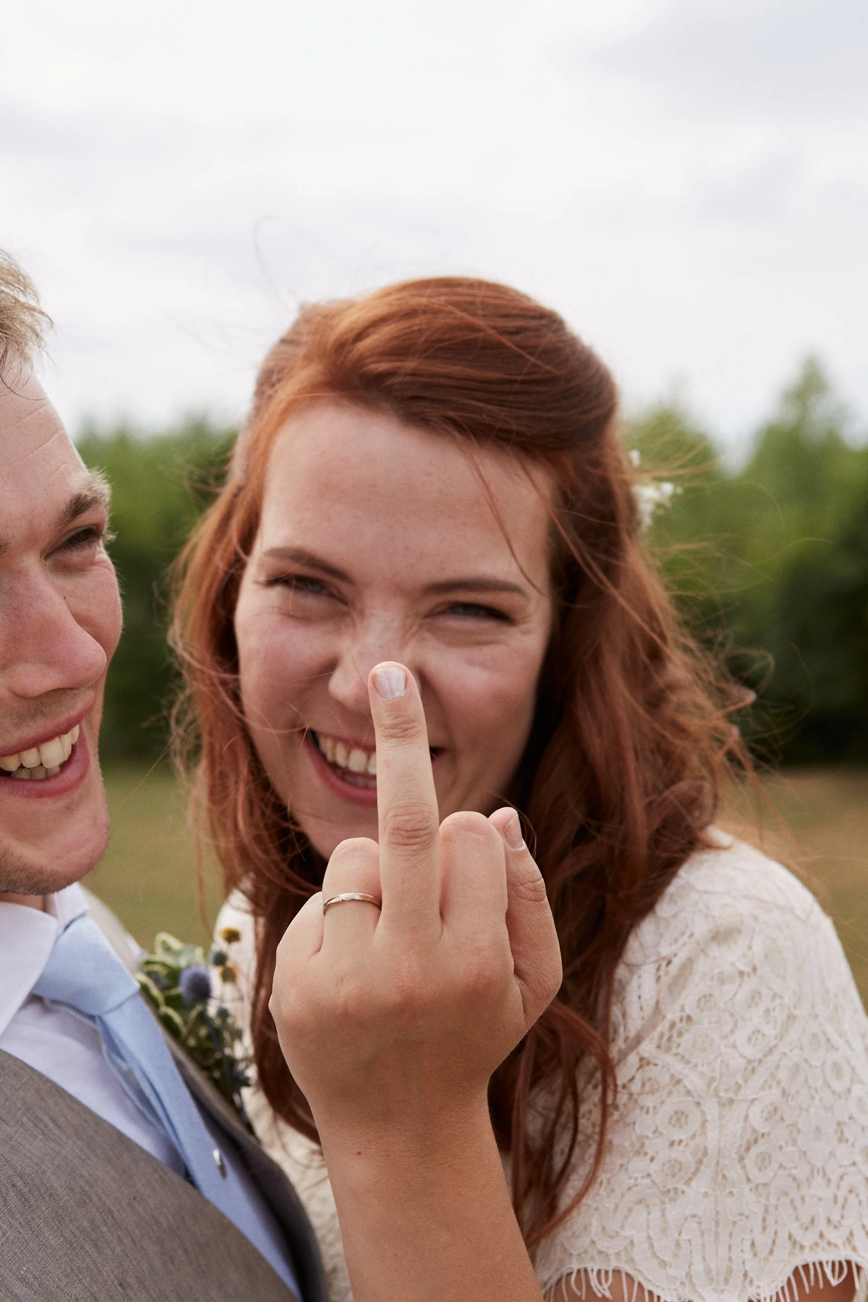 A woman with red hair and a wedding dress smiling and showing her middle finger, standing next to a man in a suit outdoors.