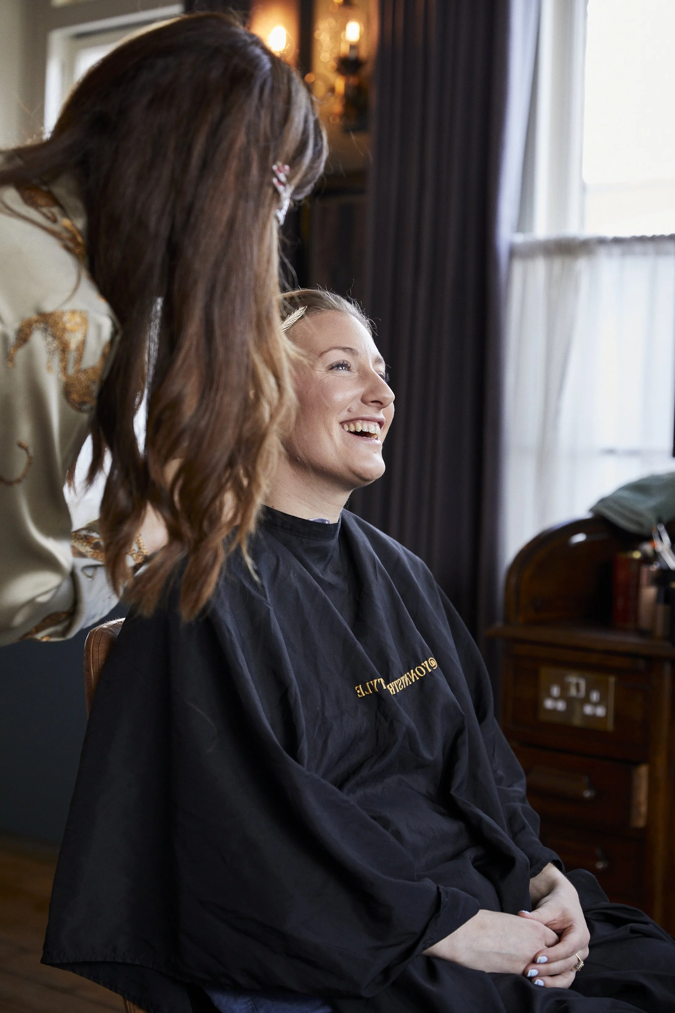 A woman is sitting in a salon chair, smiling and getting her hair styled by a hairstylist. The salon has a vintage feel with dark curtains, a wooden furniture piece, and warm lighting.