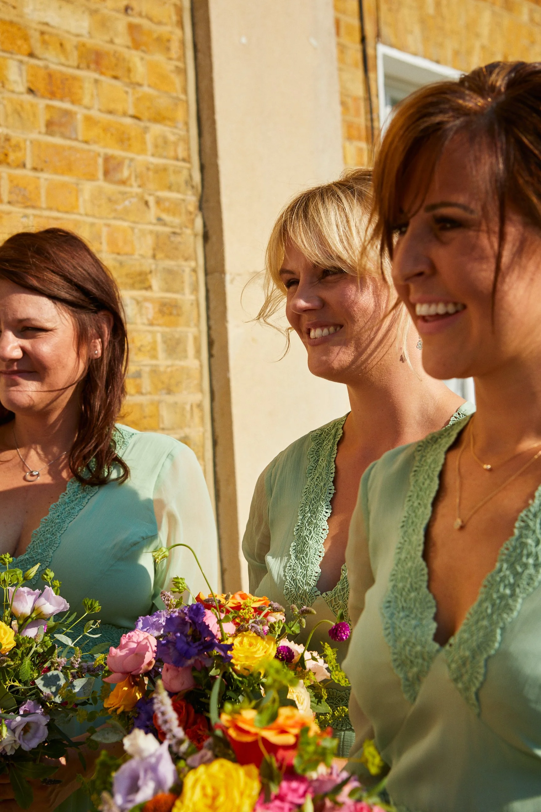 Three women smiling outdoors near a brick wall, holding a colorful bouquet of flowers, wearing pastel green dresses with lace details.