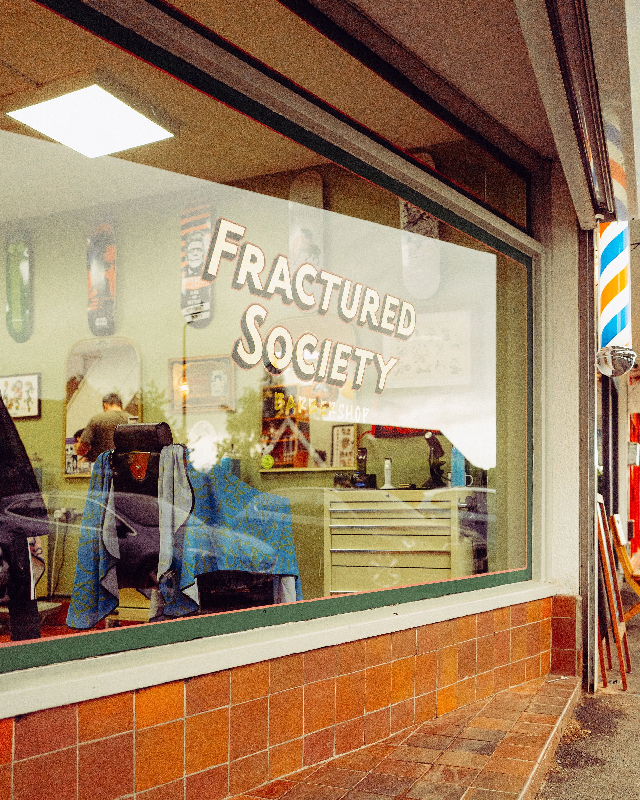 View of a storefront window with the words 'FRACTURED SOCIETY' and 'BARBERSHOP' written on it. The interior of the barbershop has chairs and posters visible through the glass. Outside, there is a sidewalk with brick tiles and part of a building with a traditional-striped barber pole near the corner.
