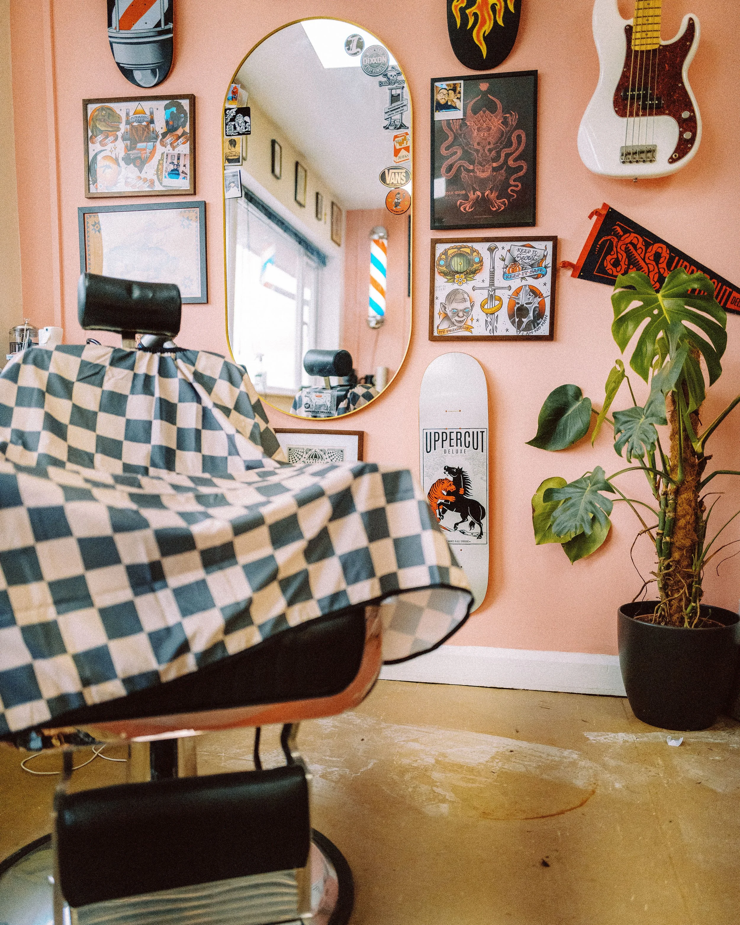 A room with pink walls decorated with various framed and unframed posters, a large mirror, a white electric guitar, and a skateboard. There is a plant with large green leaves and a checkered chair in the foreground. The floor is wooden. There are some reflections and hanging decorations visible in the mirror.