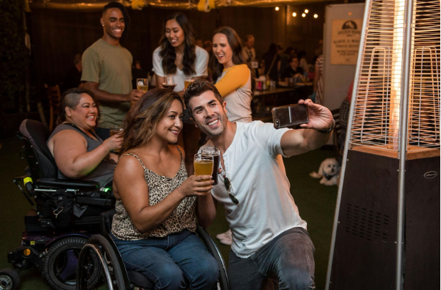 Diverse group of friends, including a wheelchair user, smiling and taking a selfie while enjoying drinks at an outdoor gathering—representing inclusive community support and the importance of social connection in recovery.