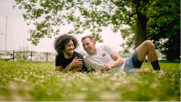 Two men, one Black and one white, lying on the grass smiling and enjoying each other's company—symbolizing LGBTQ+ joy, representation, and emotional support in healing from eating disorders.