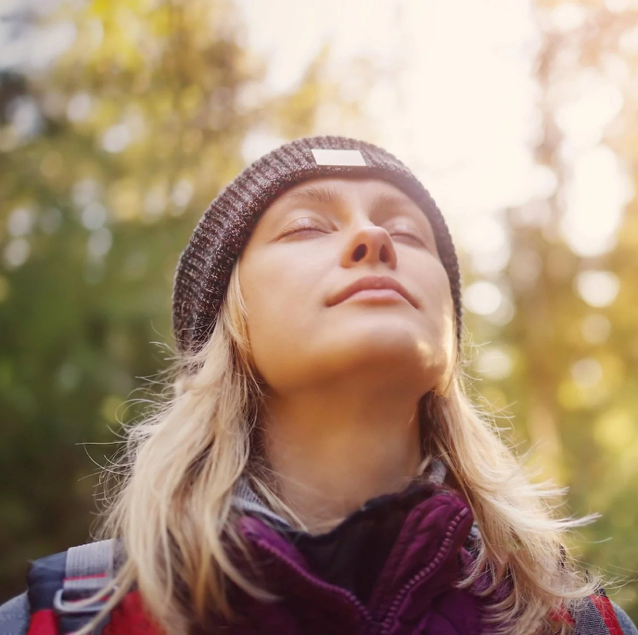 A blonde woman in a beanie closes her eyes and lifts her face toward the sun in a forest, embodying calm, breath-focused grounding—a key somatic therapy tool for easing anxiety before meals.