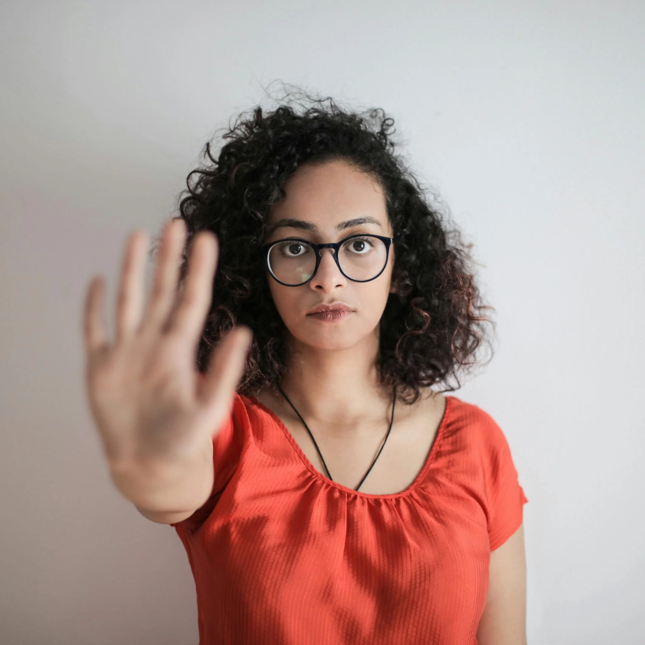 Woman with curly hair and glasses holding her hand up in a stopping gesture—representing setting boundaries, saying no to diet culture, or protecting personal healing space during recovery.