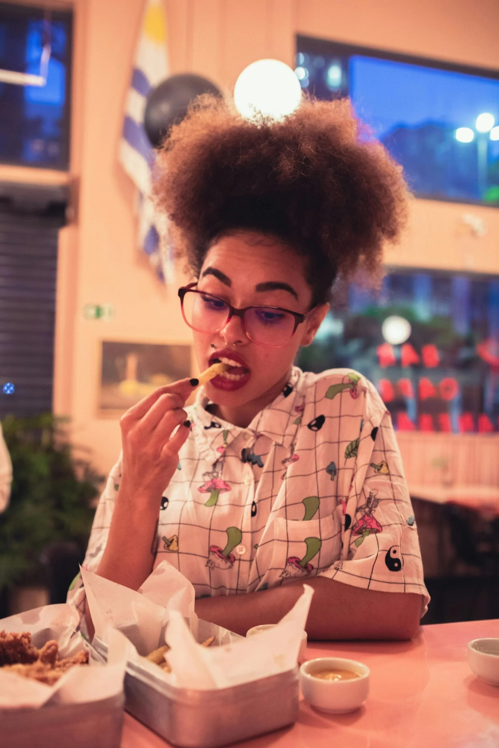 A woman wearing glasses and a playful shirt eats thoughtfully at a restaurant table, symbolizing the complexity of food-related anxiety and the journey toward intuitive, stress-free eating.