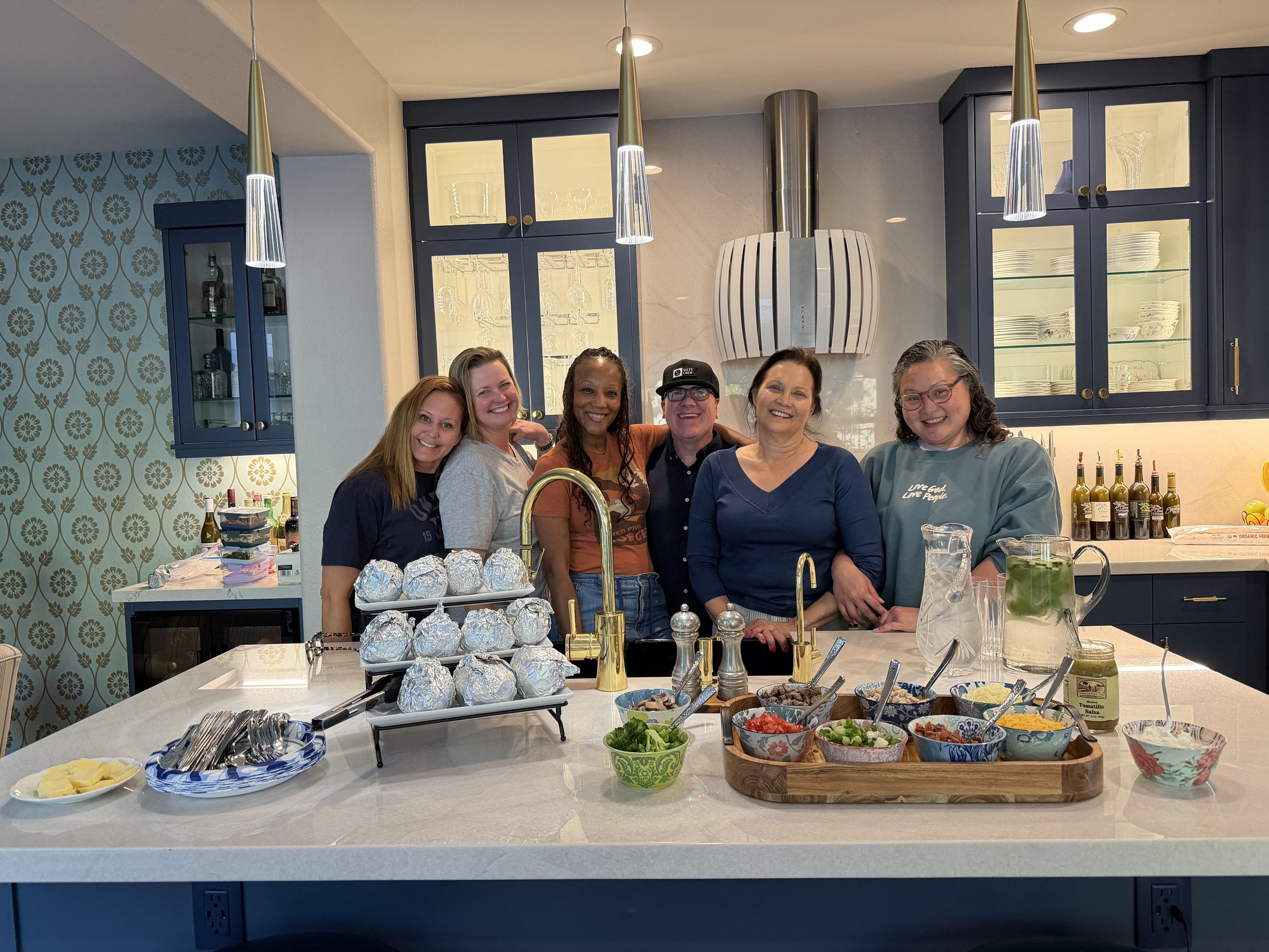 Group of six people standing in a kitchen behind a white kitchen island with food items. The kitchen features blue cabinets with glass doors and modern lighting. There are various bowls of salsa, chopped vegetables, and foil-wrapped dishes on the island.