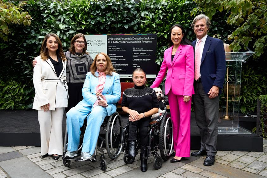A group of six people is standing and sitting for a photo outdoors. Two women in wheelchairs