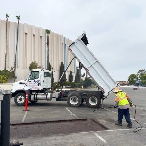  A rectangular area is cut out of an asphalt parking lot. A worker is near the hole and there is a dump truck in the background. 