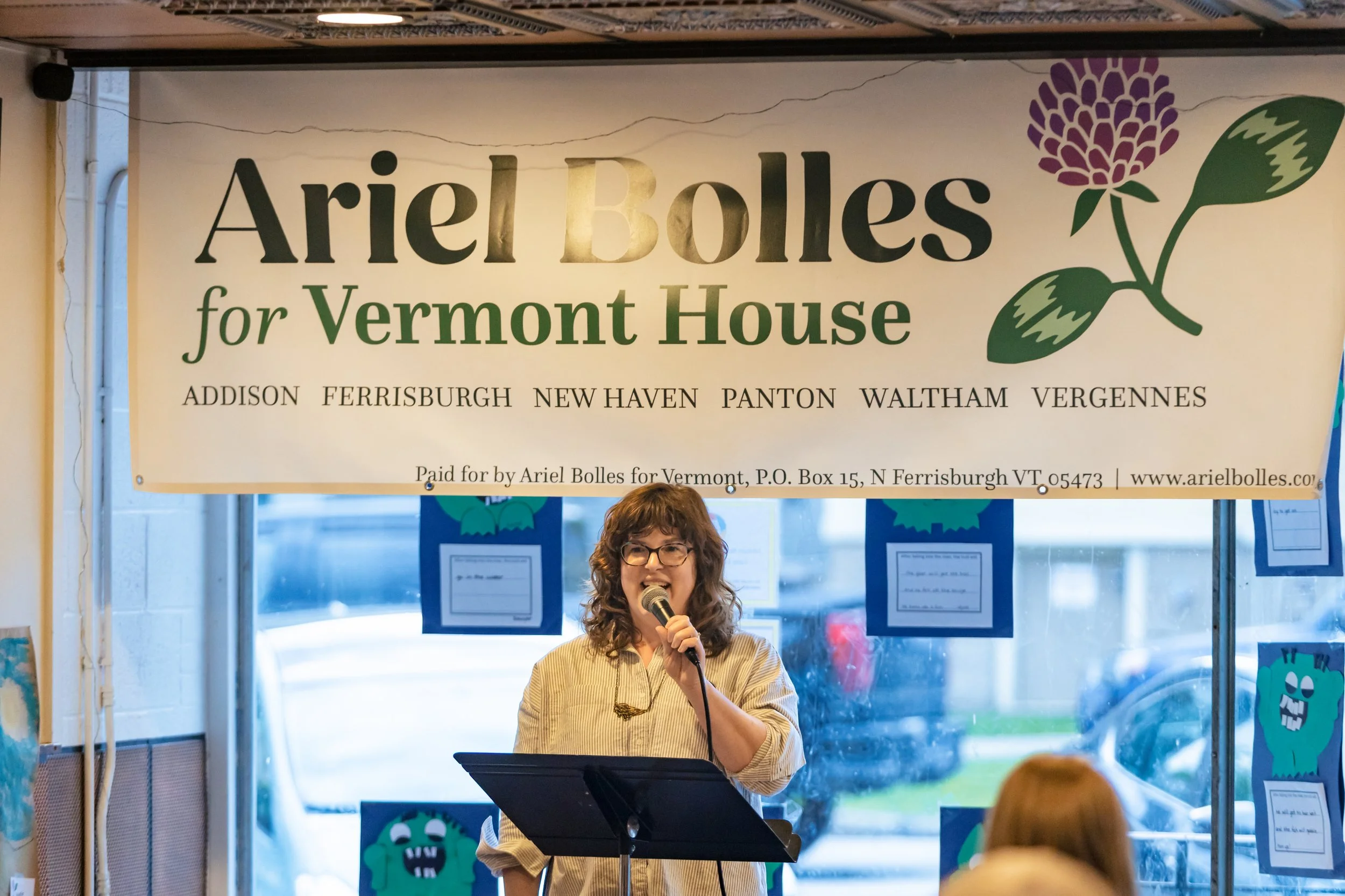 Ariel stands with a microphone in front of a banner that reads: "Ariel Bolles for Vermont House, Addison, Ferrisburgh, New Haven, Panton, Waltham, Vergennes, Paid for by Ariel Bolles, P.O. Box 15, N Ferrisburgh VT 05473, www.arielbolles.com"