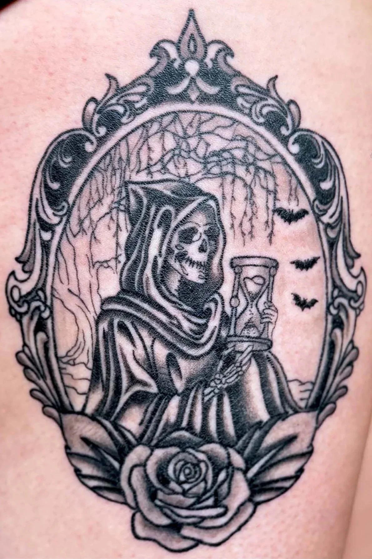 An illustrative black and gray tattoo of the Grim Reaper holding an hourglass, framed by an ornate vintage border with bats and a rose.