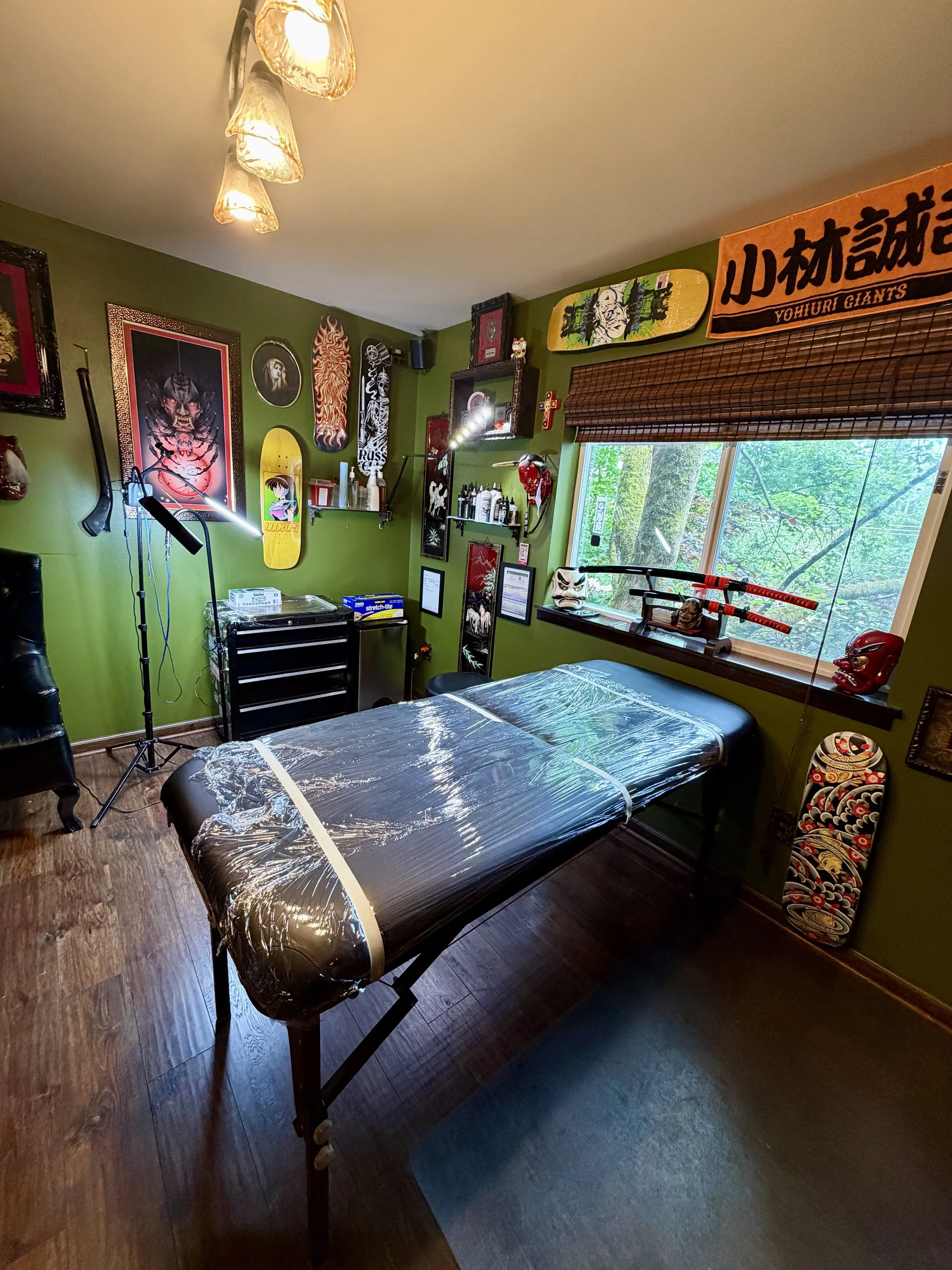 A professional hydraulic tattoo table wrapped in protective plastic inside a private studio, with katana swords displayed on a window sill and skateboard deck art on olive green walls.