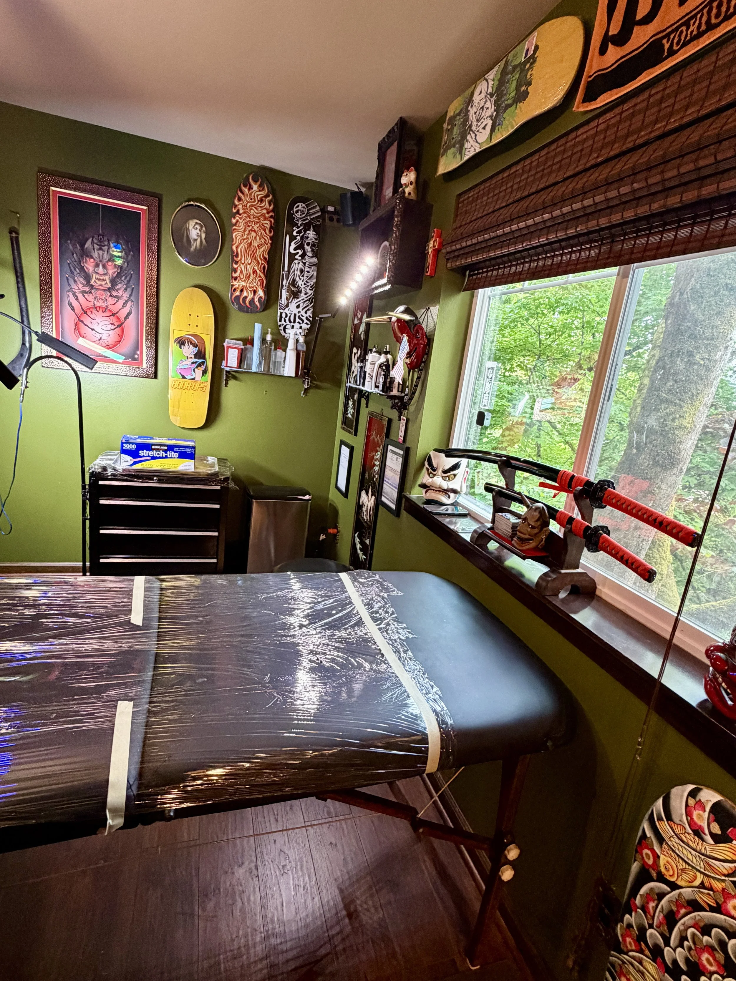 Close-up of a clean, plastic-wrapped tattoo table next to a window overlooking trees, inside a private tattoo studio with custom artwork and professional lighting.