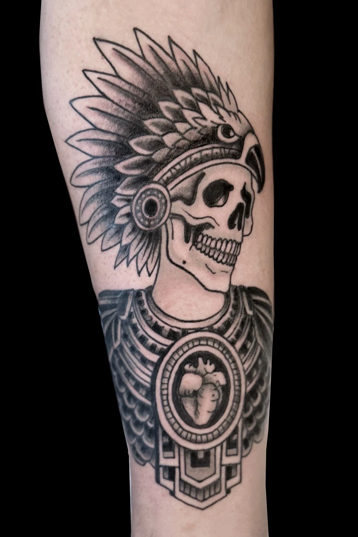A bold black and gray tattoo showcasing a human skull wearing a highly detailed, traditional Native American feathered headdress.