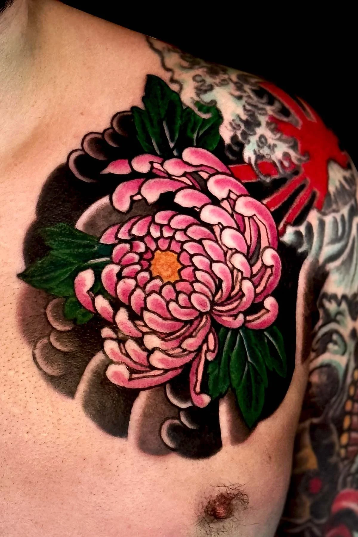 Close-up of a shoulder tattoo featuring a pink chrysanthemum flower with green leaves, outlined in black, with a black background shading.