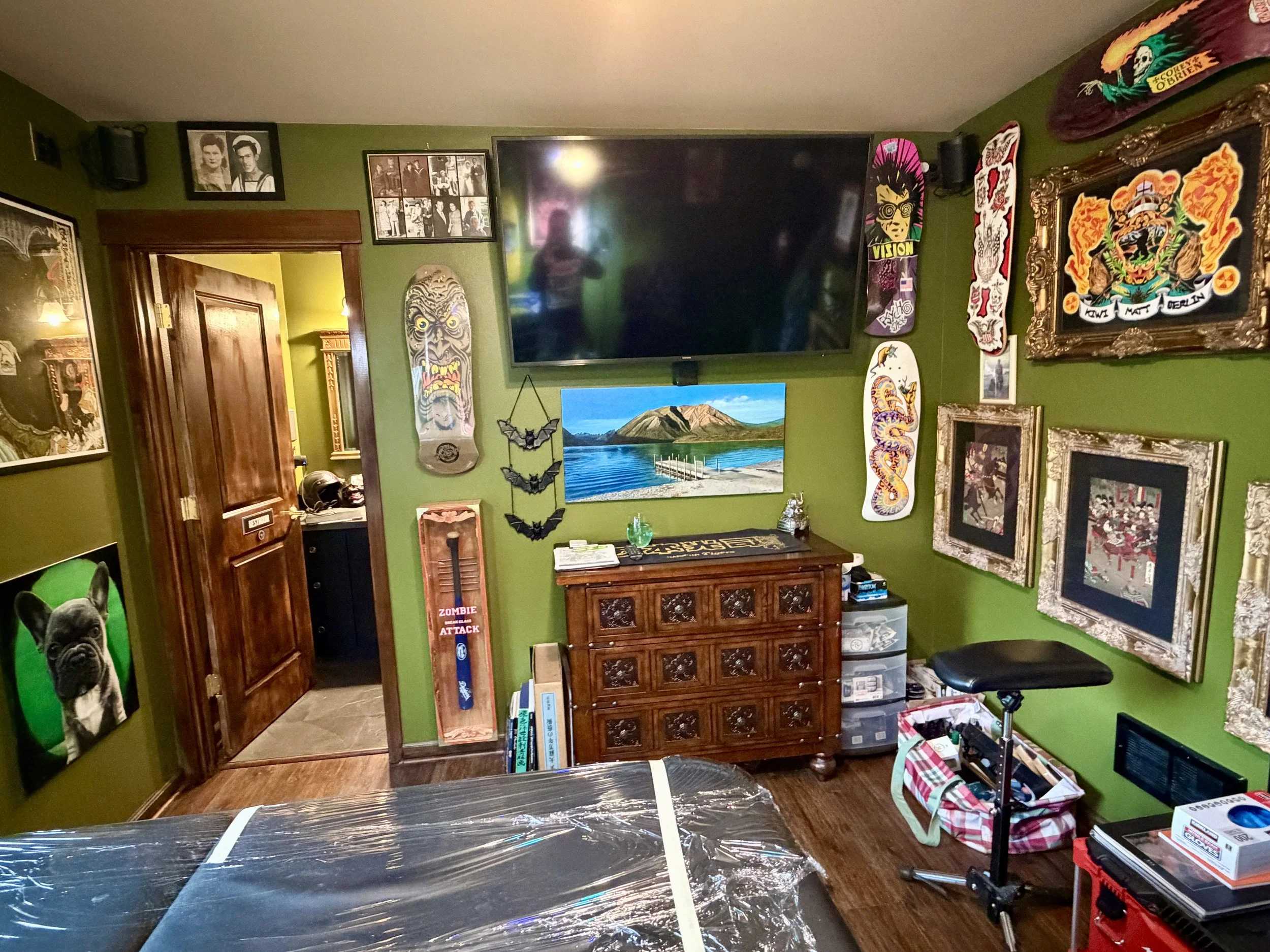 Private tattoo studio client comfort area featuring a wall-mounted flat-screen TV, a vintage wooden dresser, smart-home lighting, and framed tattoo flash art.