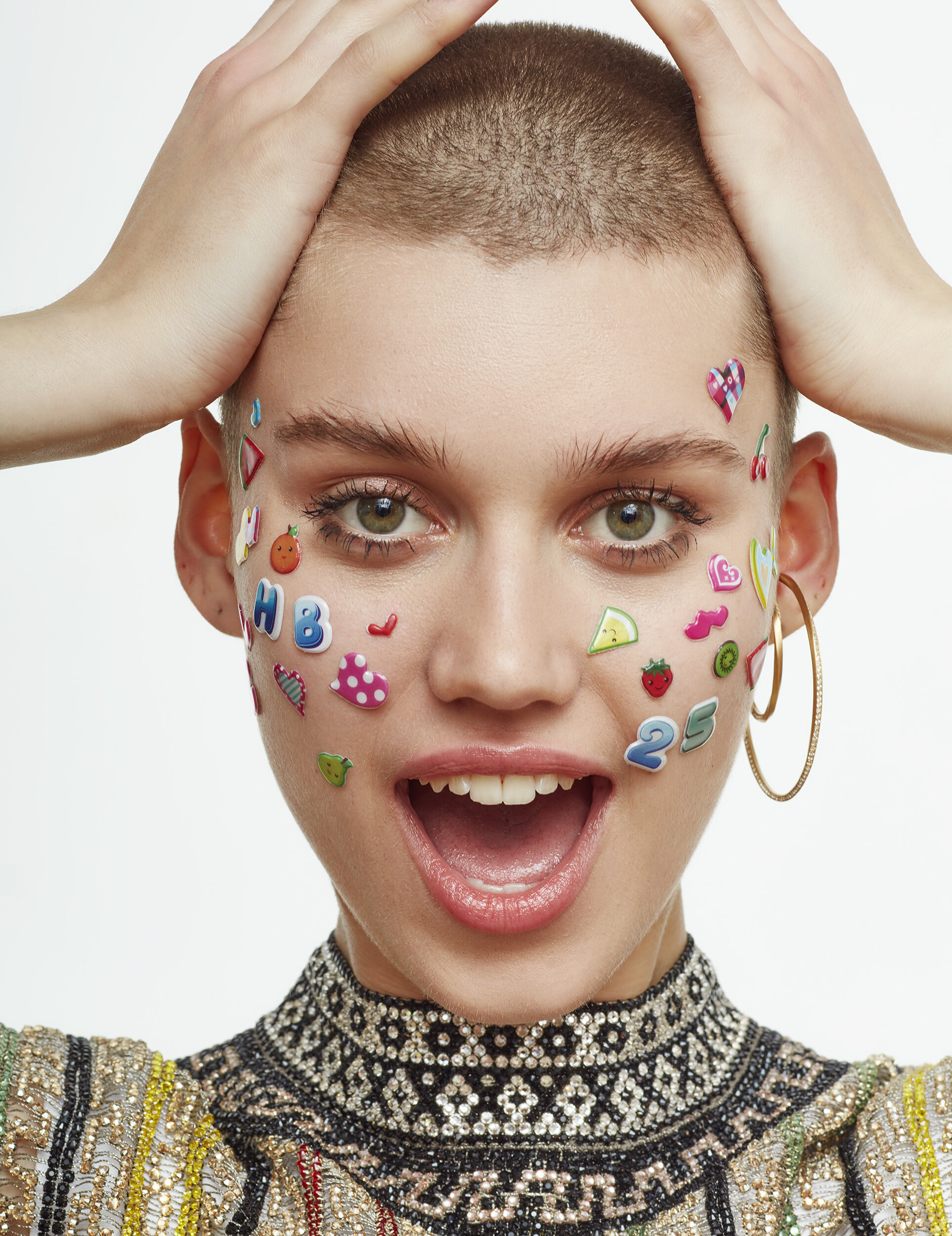 A woman with a shaved head has colorful stickers on her face, including letters, numbers, and images of fruits, with her hands on her head, smiling with an open mouth, wearing a beaded top and a large hoop earring.