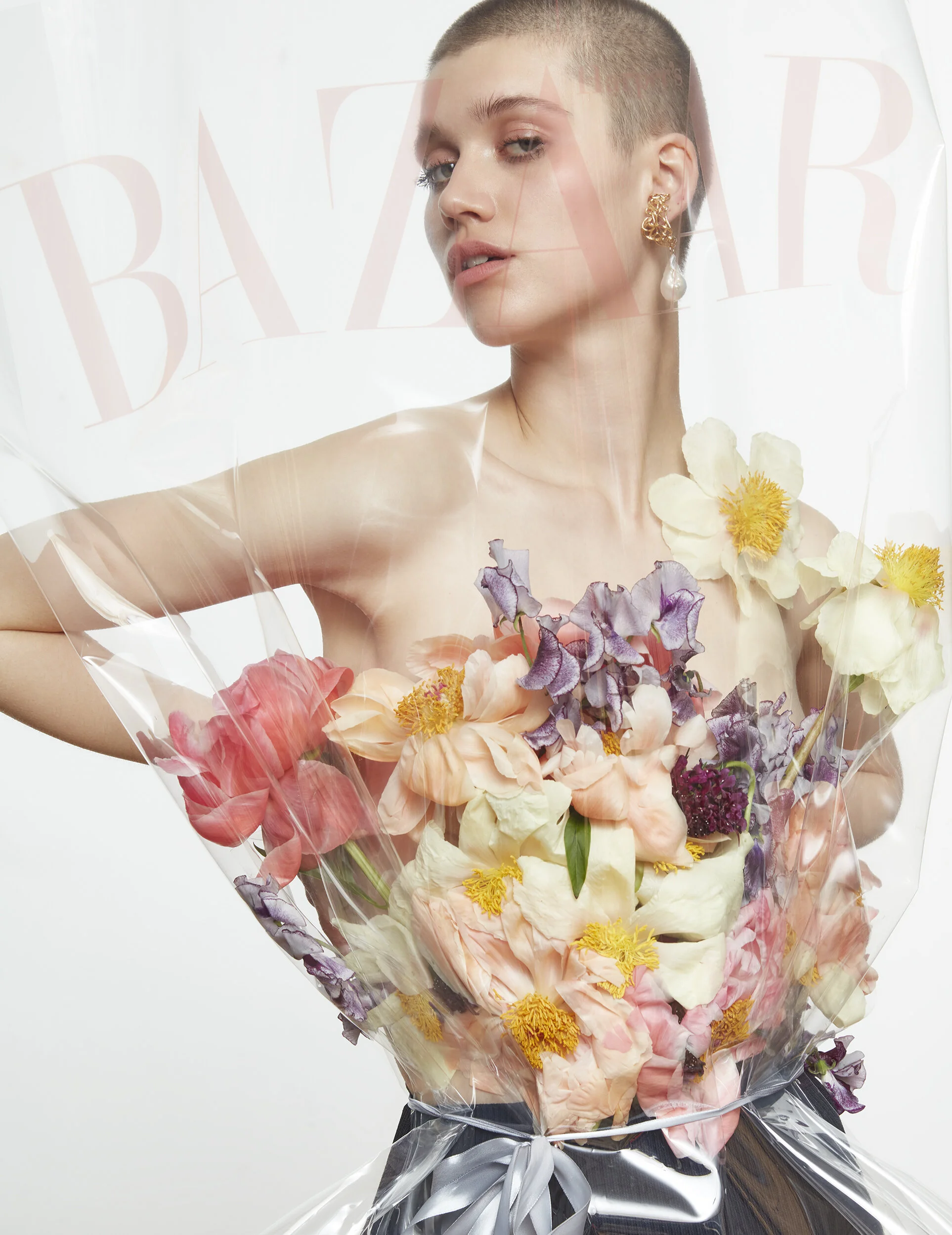 A woman with short hair and earrings holds a bouquet of various colorful flowers wrapped in transparent plastic and tied with a silver ribbon, with the word 'BAZAAR' visible behind her.