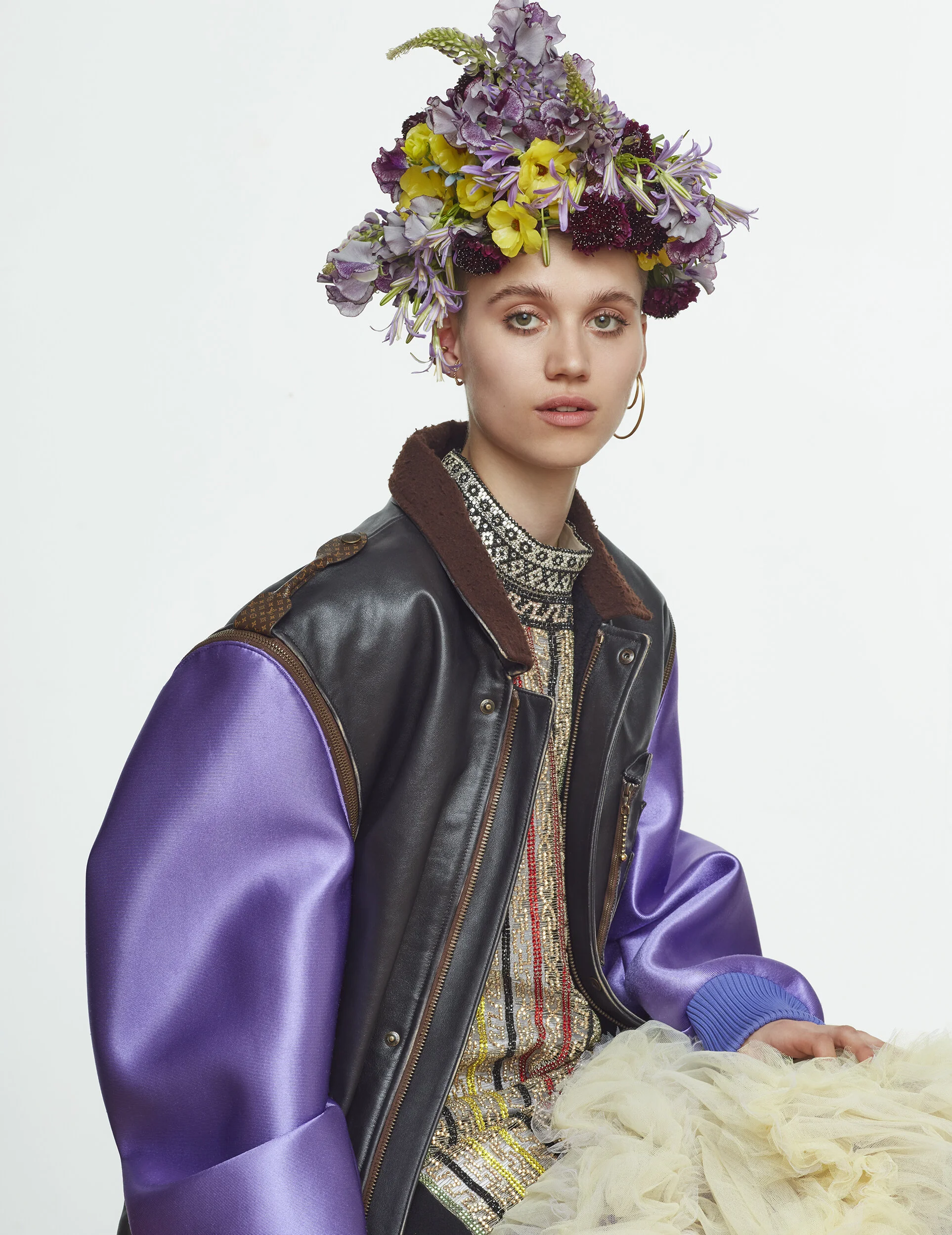 A woman with a floral headdress, wearing a purple and black jacket and sitting with a cream-colored tulle fabric.