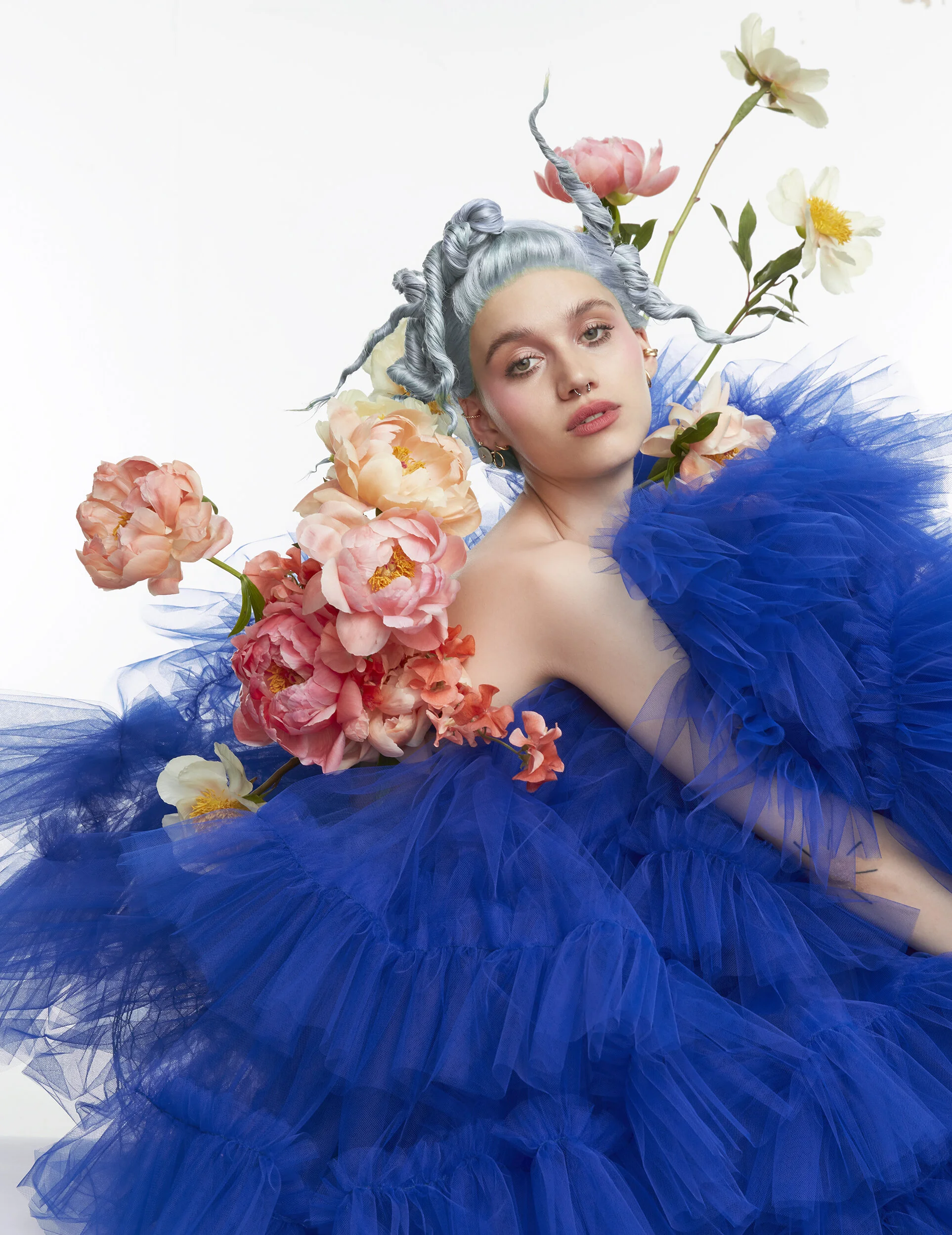 A woman with styled gray hair adorned with flowers, wearing a voluminous royal blue tulle gown, surrounded by pink and white flowers against a plain white background.