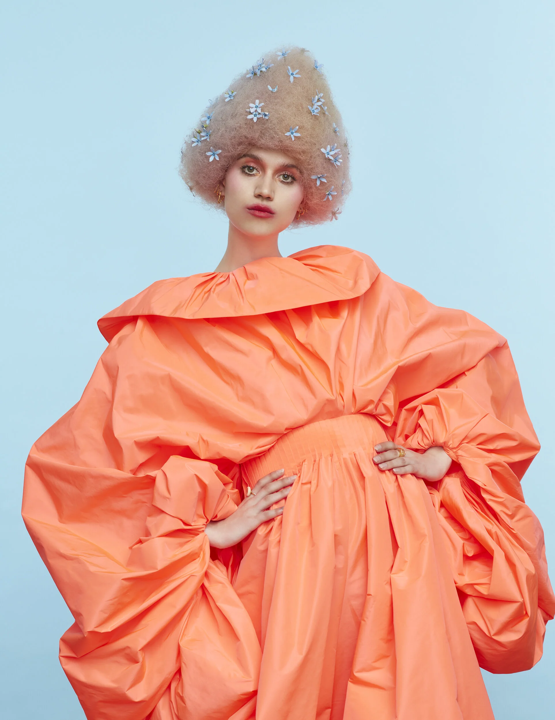 A woman with curly blonde hair styled with small blue flowers, wearing a voluminous, bright orange dress with puffy sleeves, standing against a light blue background.