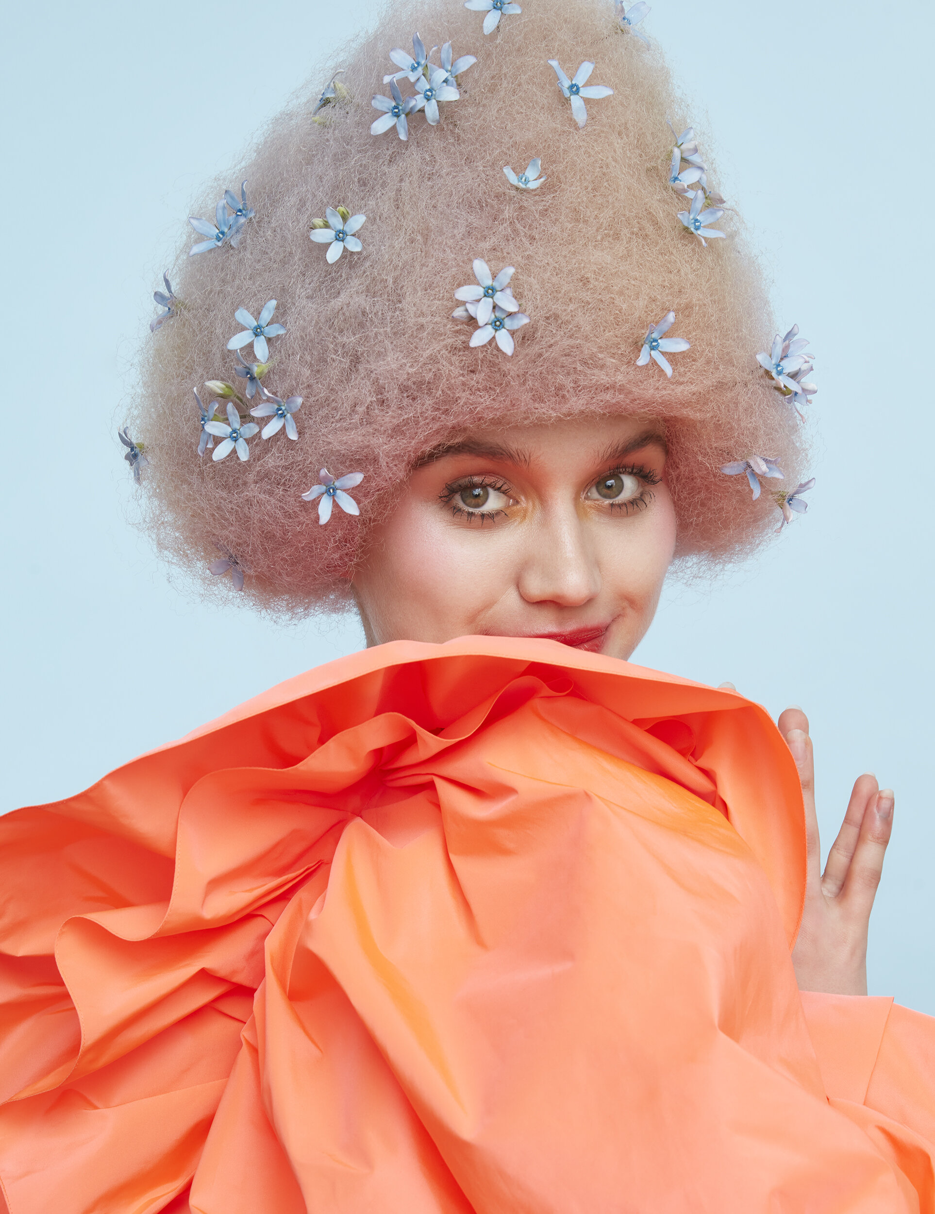 A woman with curly pink hair adorned with small blue and white flowers, holding an orange fabric with ruffles.